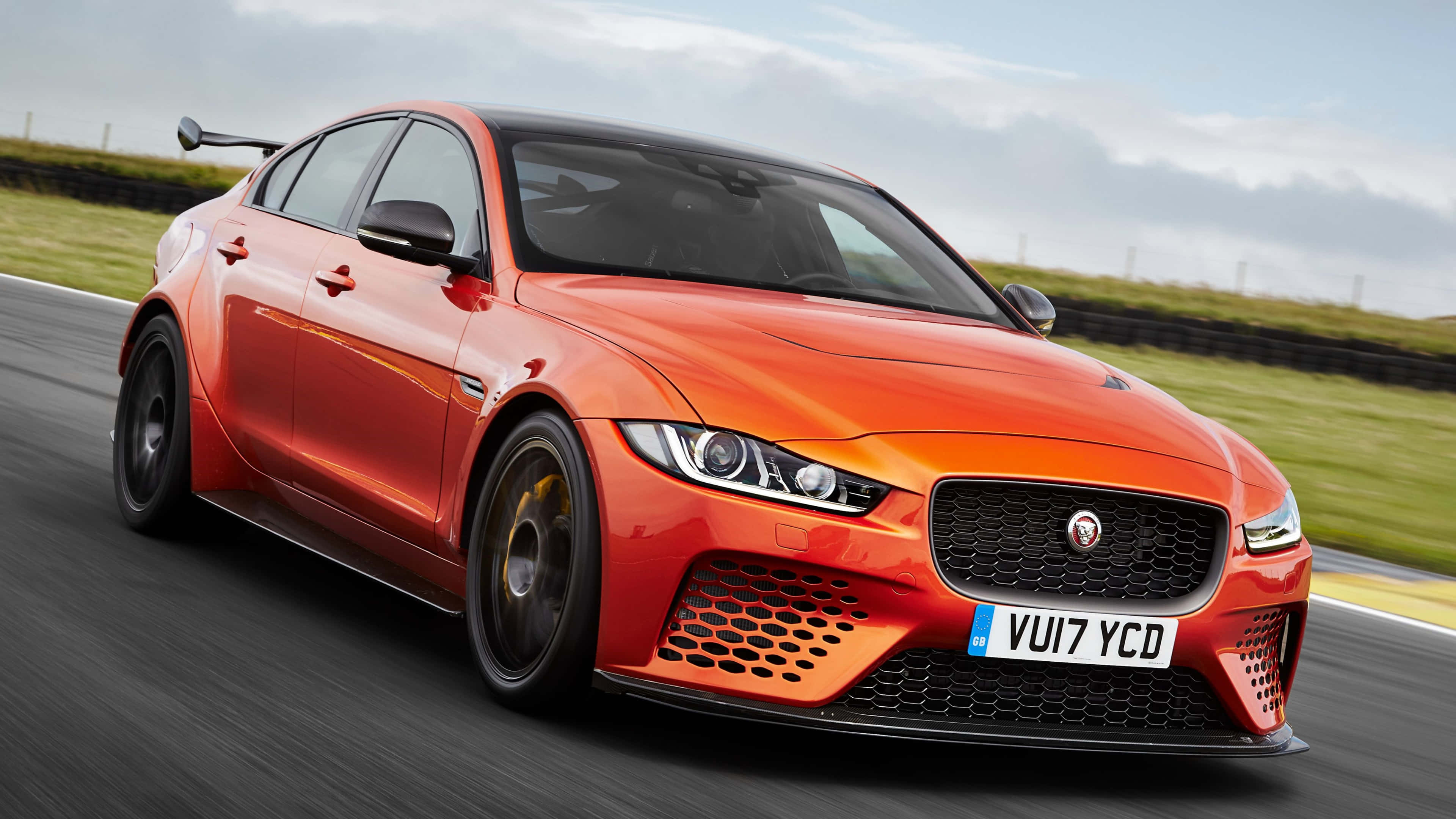 4K Project Cars 橙色捷豹XE SV背景 壁紙 — 一輛充滿活力的橙色Jaguar XE SV Project 8在賽道上疾馳，背景是模糊的綠色草地和多雲的天空。