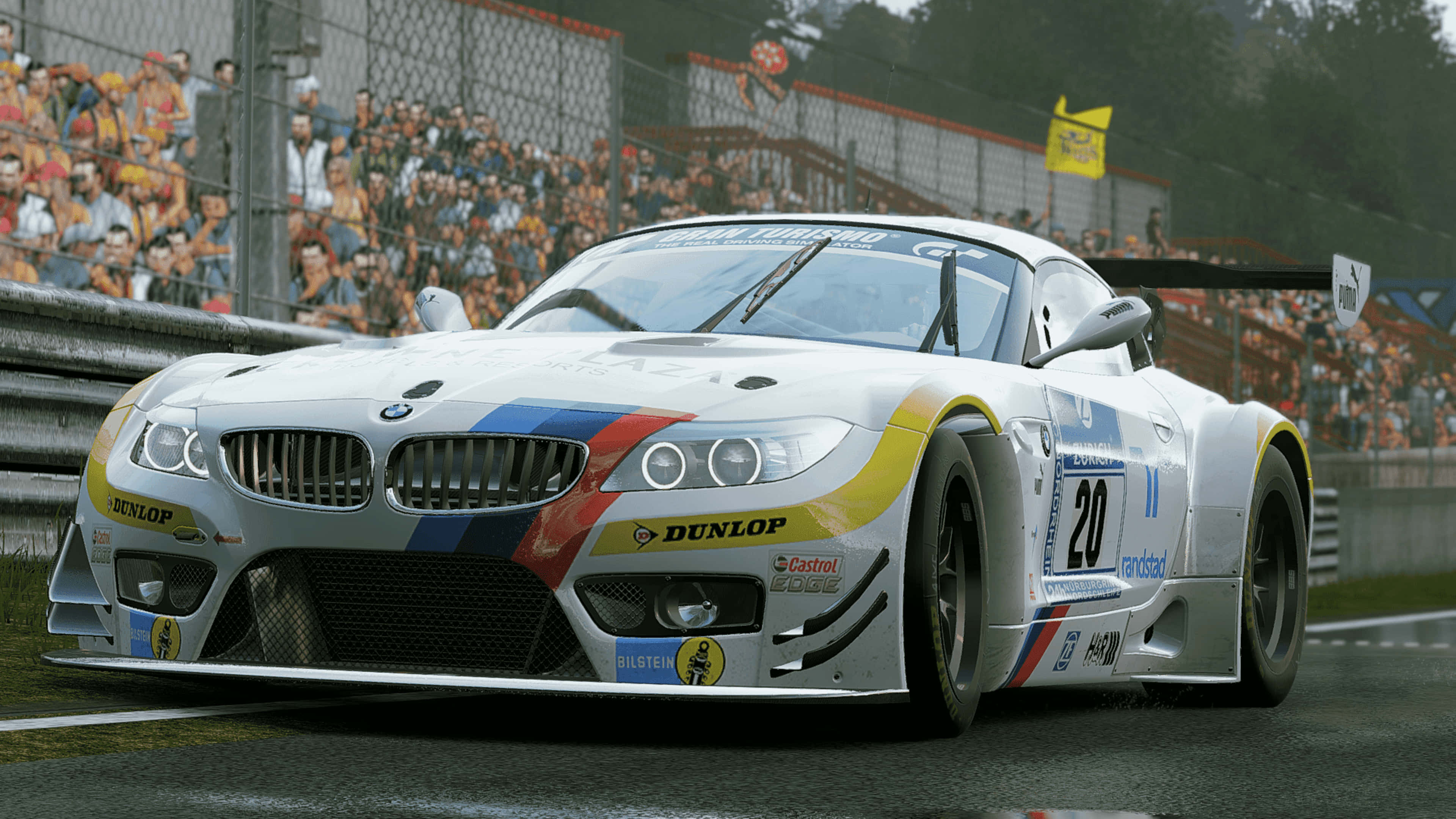 4K Project Cars 白色 BMW Z4 GT3 (E89) 背景 壁紙 — 一輛白色BMW賽車，帶有藍、紅、黃色塗裝，在潮濕的賽道上疾馳，圍欄後有歡呼的觀眾。
