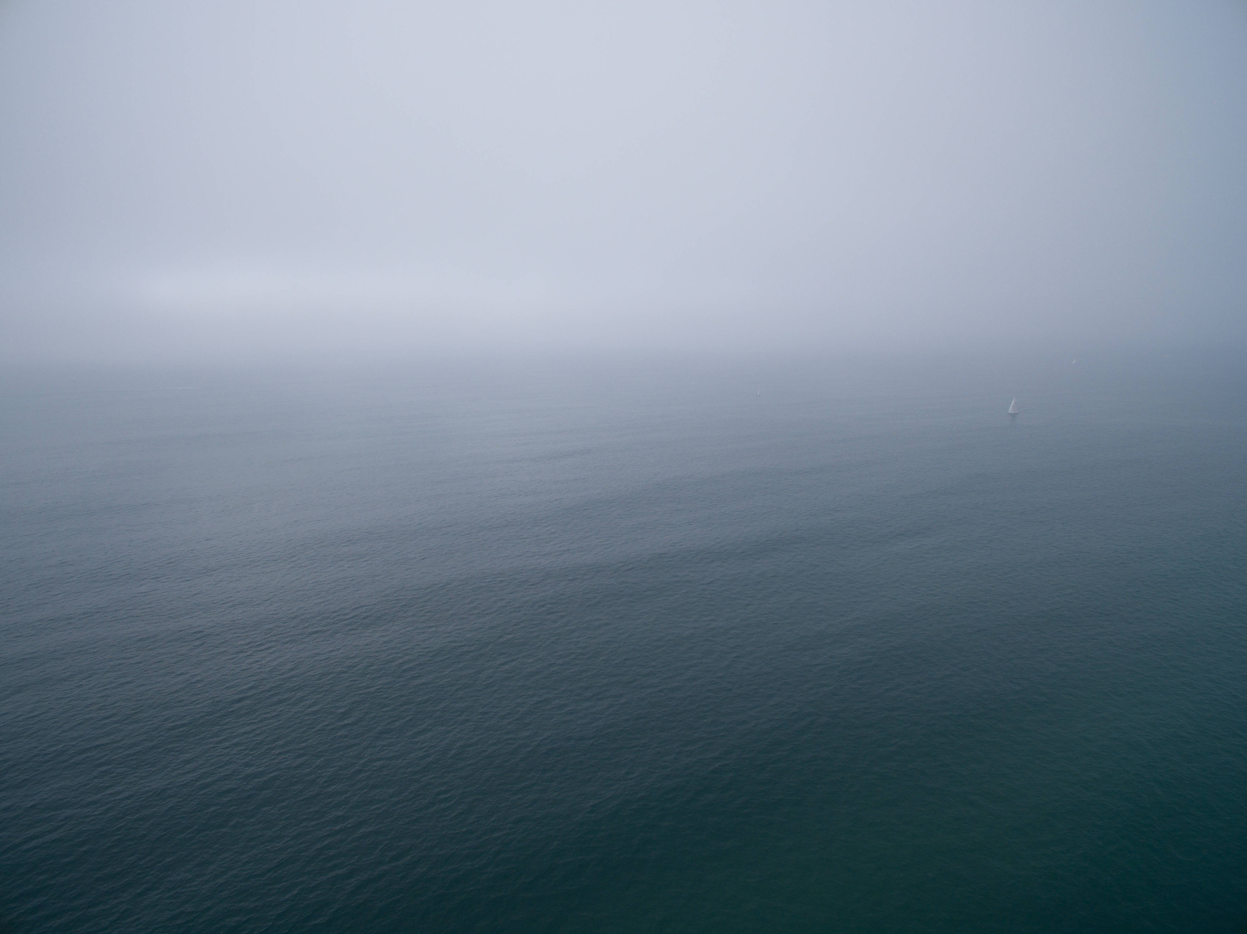 4K 海灘水景 壁紙 — 一艘孤獨的帆船在灰色天空下穿過霧氣瀰漫的海面。