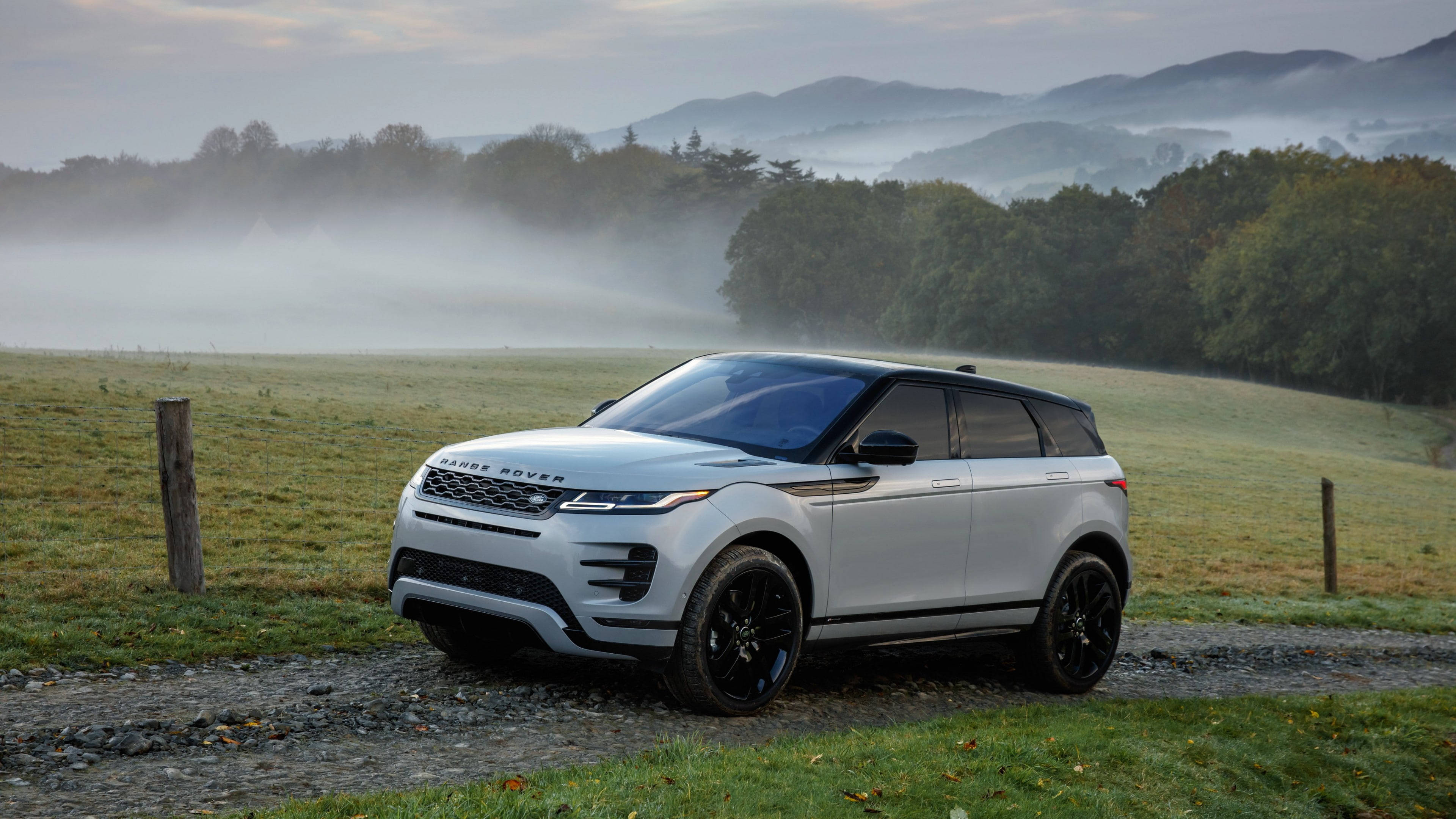 高質感 4K Range Rover 駛過鄉野風光 壁紙 — 一架時尚的銀色Range Rover Evoque停泊在霧氣瀰漫的碎石路上，背景是起伏的綠色山丘和霧濛濛的森林。