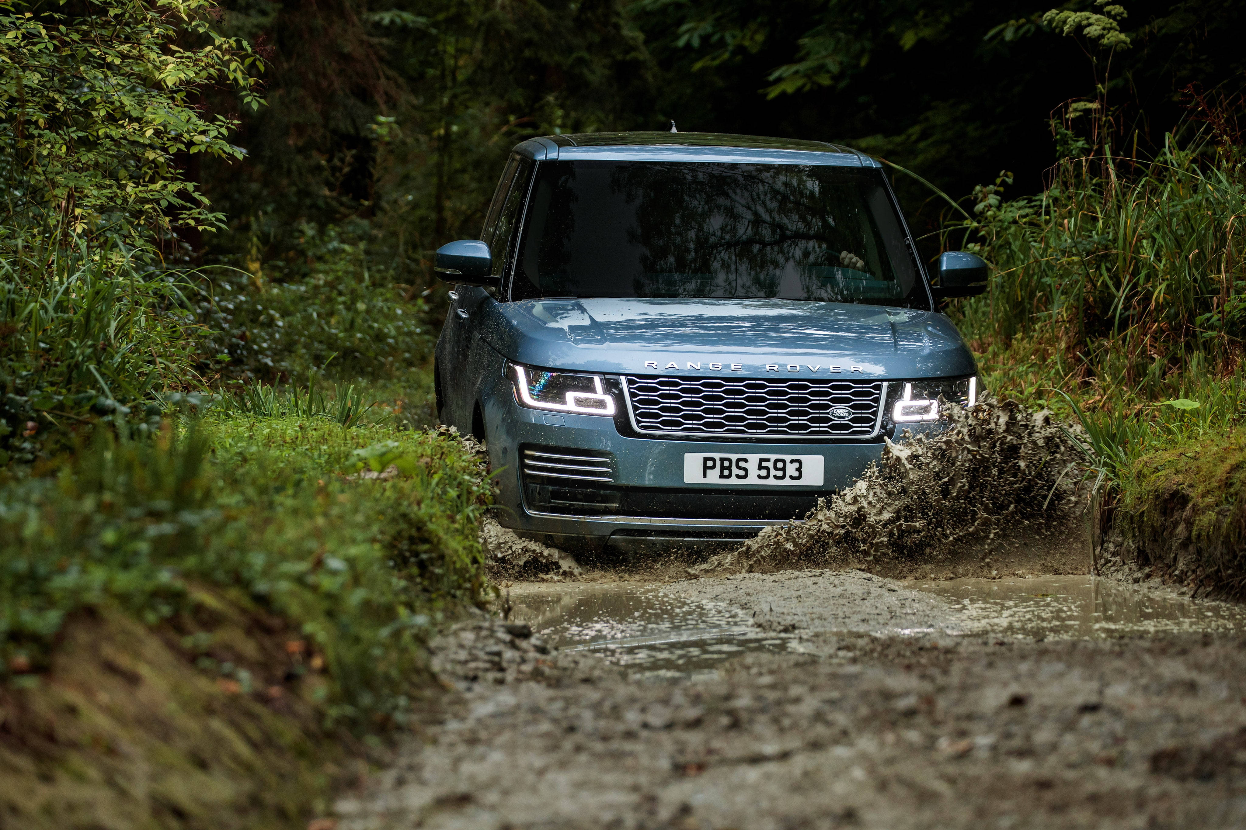 粗獷中的優雅 - 4K Range Rover 穿越多樣地形 壁紙 — 一輛時尚的藍色Range Rover在泥濘的森林小徑上行駛，車頭燈亮起，水花四濺。