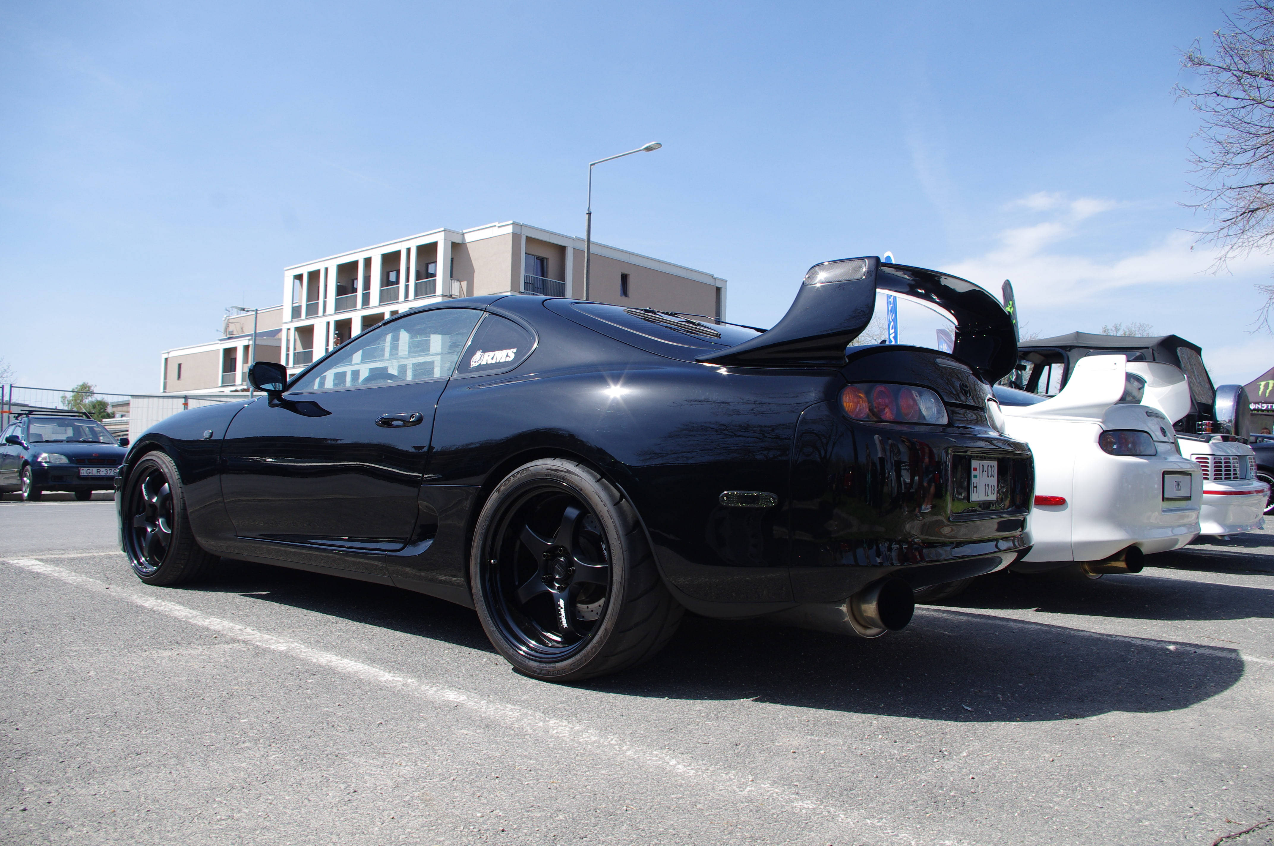 4K Supra 後視特寫 壁紙 — 一架亮黑色豐田Supra，尾翼升起，停泊在陽光明媚的城市空地，旁邊有一輛白色跑車。
