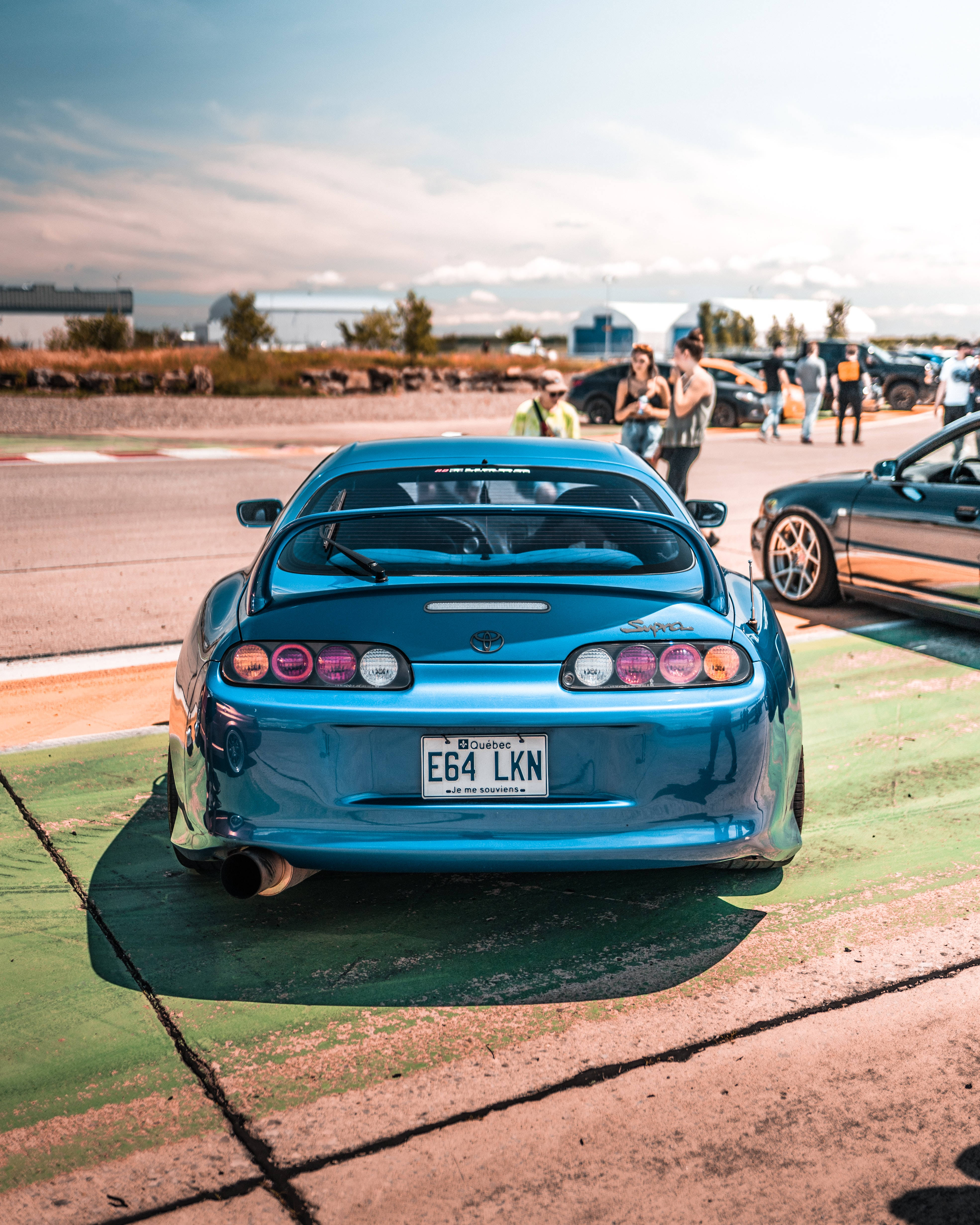 4K 丰田Supra 肯尼亞藍黑配色 壁紙 — 一架充滿活力的藍色跑車，配備後擾流板和定制排氣管，停在晴朗天空下的賽道旁停車場。