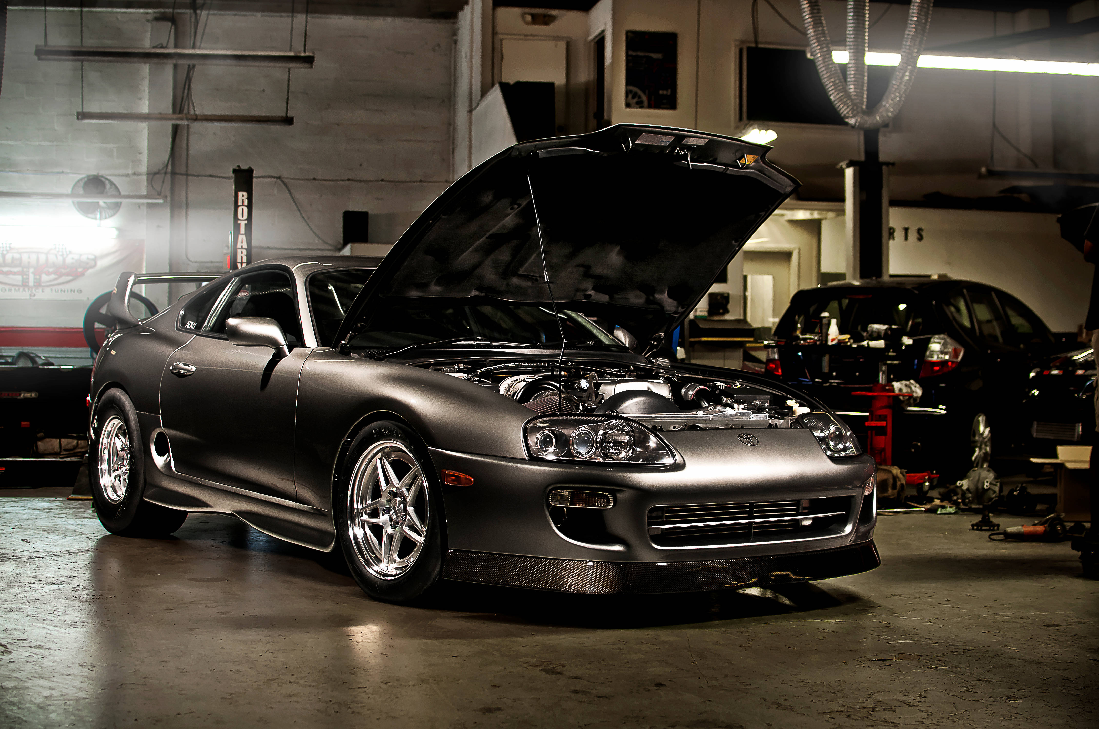 4K Supra 與城市夜景的璀璨對比 壁紙 — 一架啞光灰色豐田Supra打開引擎蓋，停放在光線昏暗的車庫中，周圍有工具和另一輛車。