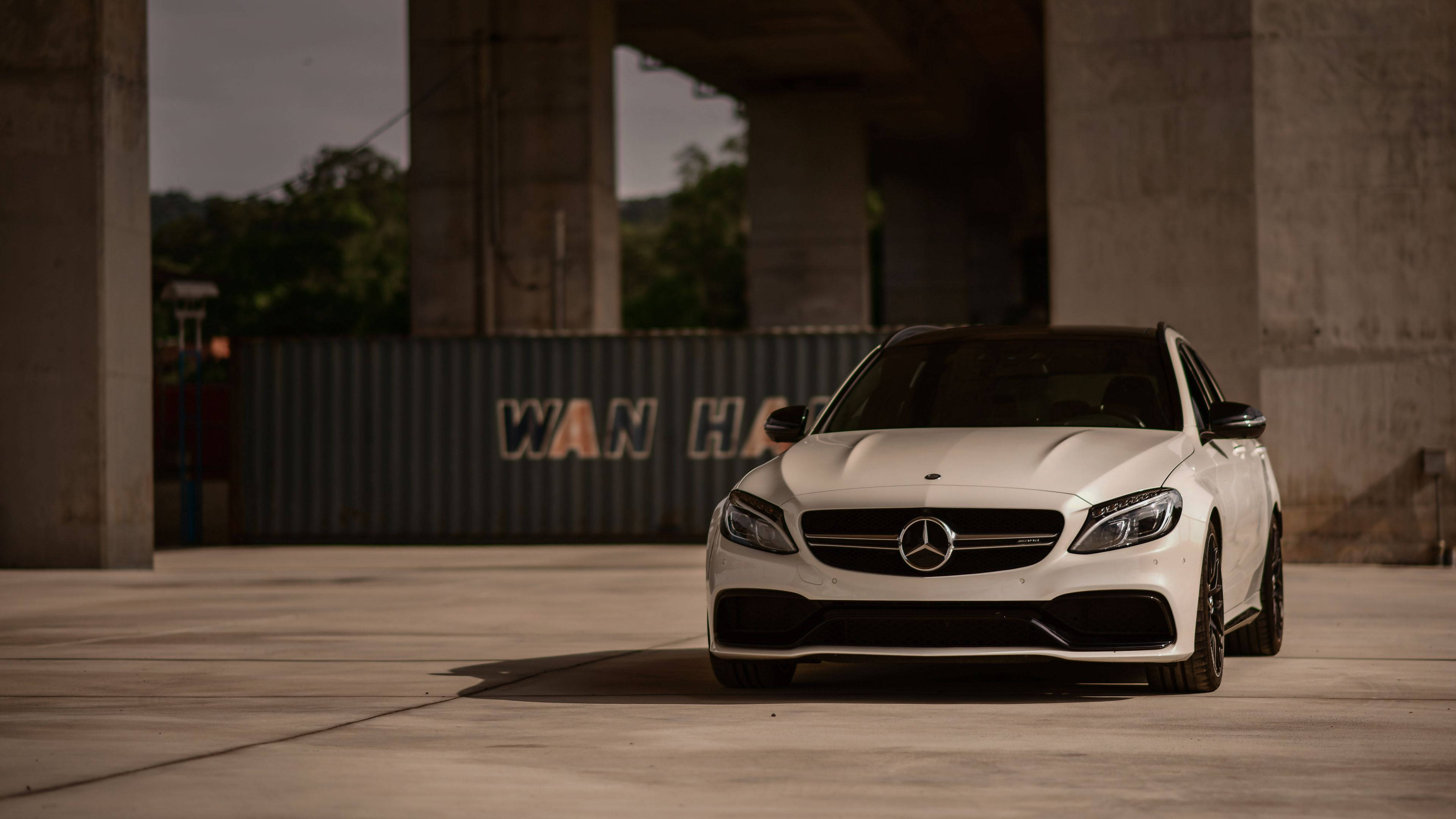 4K 梅賽德斯-奔馳白色 SUV 壁紙 — 一架時尚的白色 Mercedes-AMG C63 停泊在混凝土高架橋下，背景有工業貨櫃。