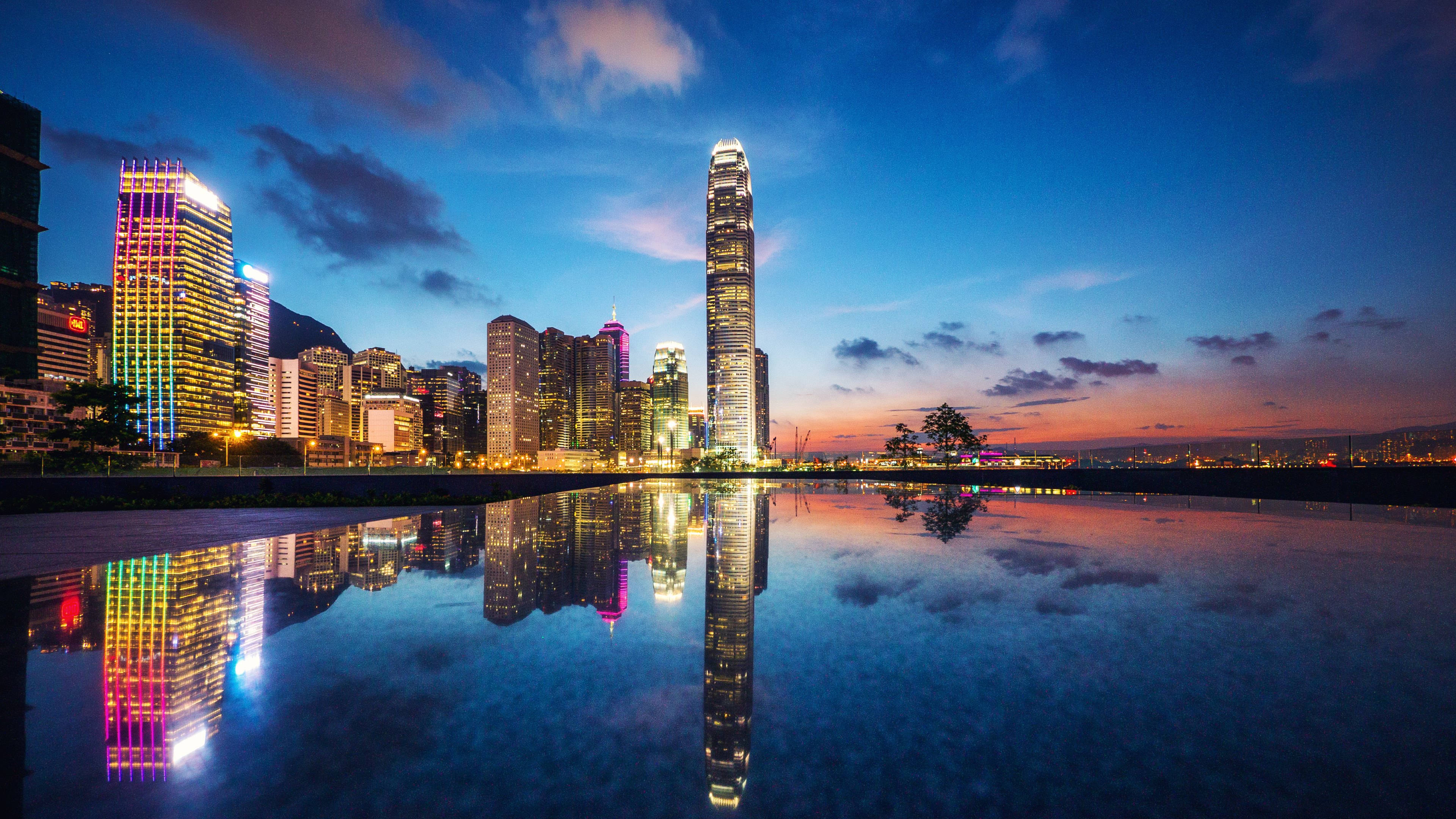 4K 超高清香港灣區城市 壁紙 — 令人驚嘆的黃昏城市景觀，摩天大樓在平靜的水面上倒映，天空呈現鮮豔的藍色和橙色。