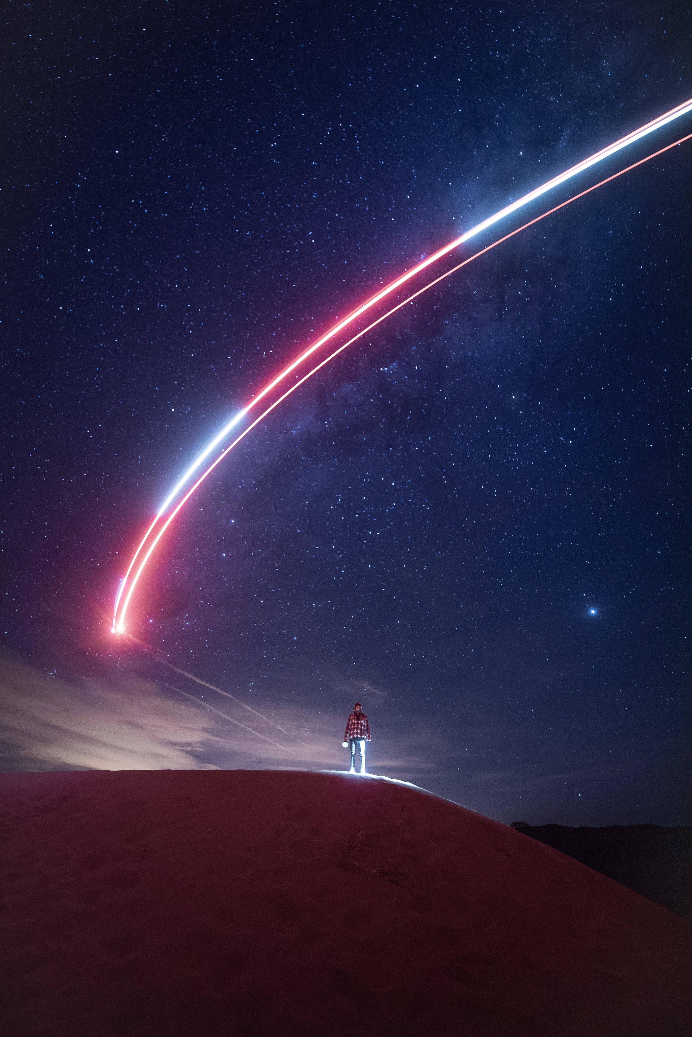 4K霓虹夜空 壁紙 — 一個孤獨的身影站在沙漠沙丘頂上，頭頂上是繁星點點的夜空，鮮紅和藍色的光跡劃過天際。