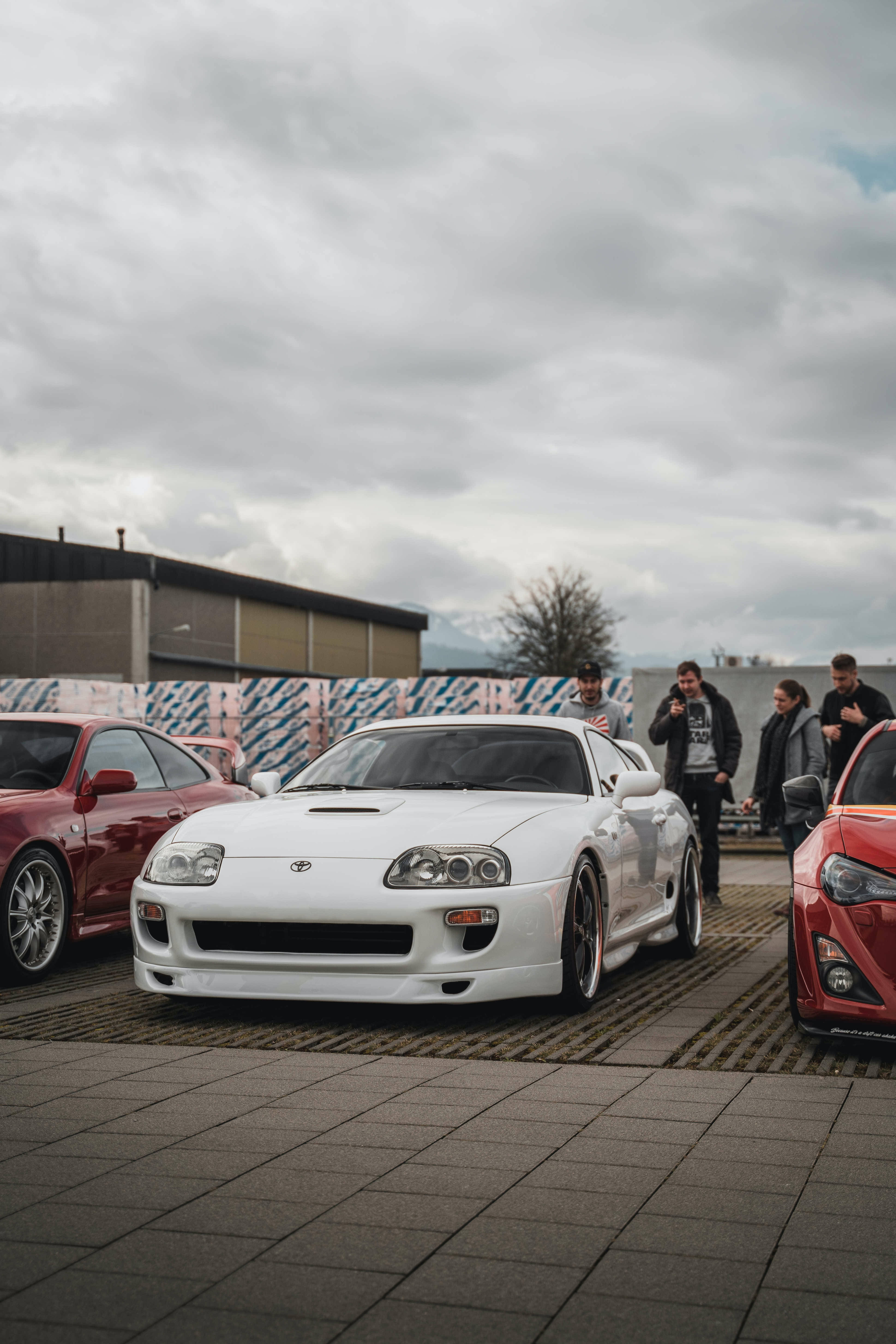 4K 《Toyota Supra》白色車款停在磚砌停車場 壁紙 — 一輛白色豐田Supra跑車停泊在瓷磚地上，人們在陰天欣賞它。