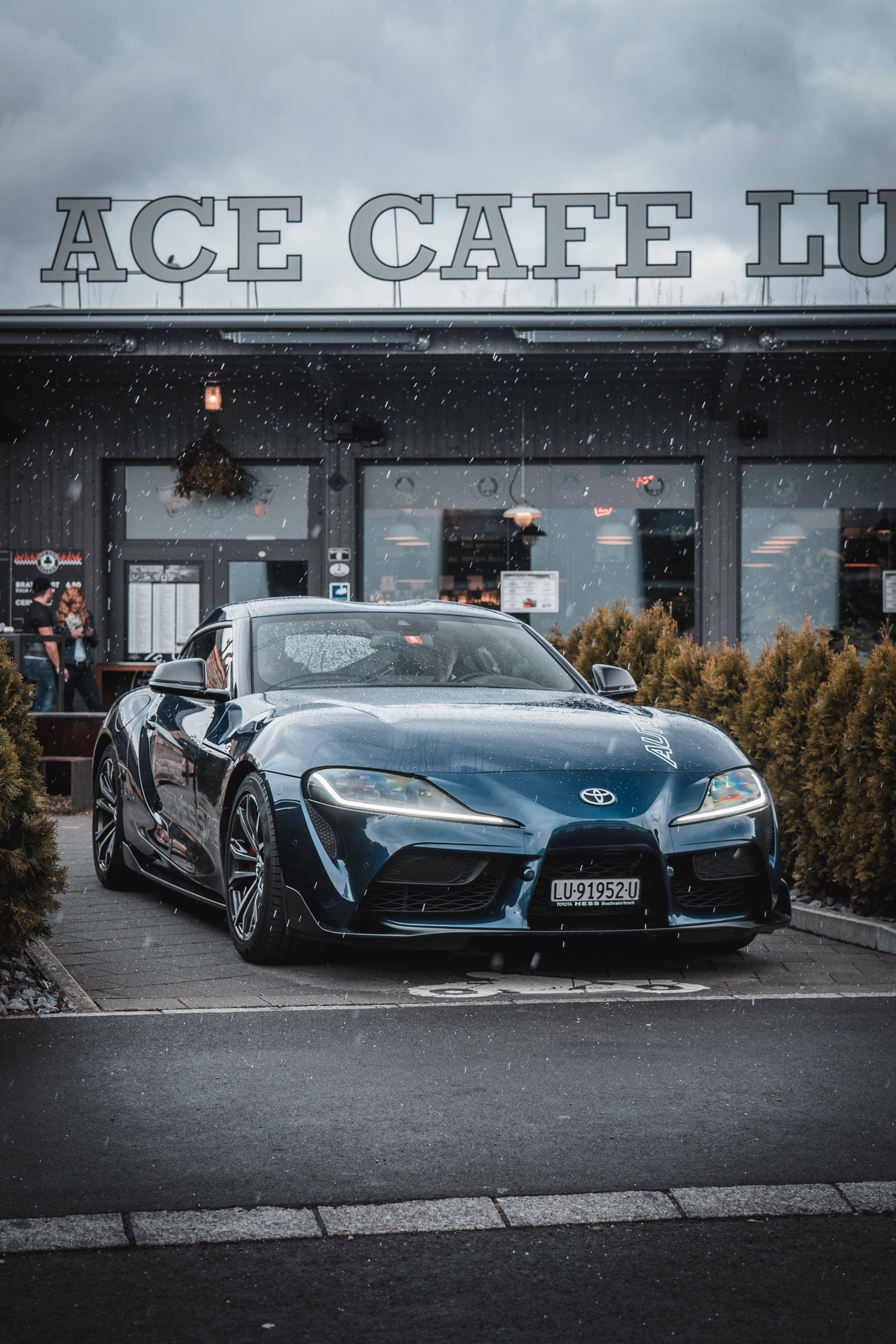 4K 《Toyota Supra》深藍色車款 壁紙 — 一架時尚的藍色跑車停泊在ACE CAFE外，天空灰暗，雪花飄落。
