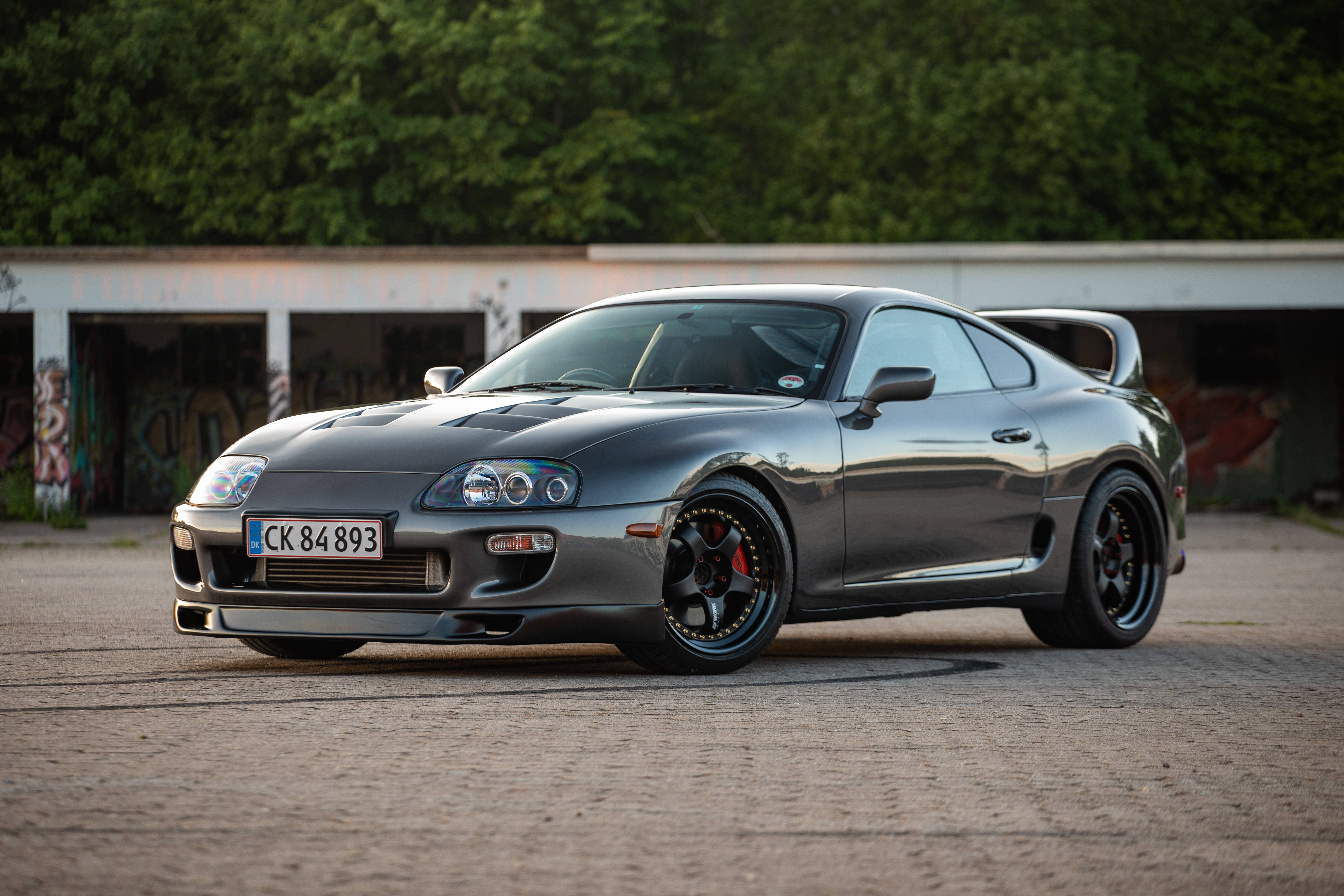 4K 《Toyota Supra》深灰車款 壁紙 — 一輛時尚的灰色豐田Supra跑車，配備黑色輪圈和紅色制動卡鉗，停在鋪砌的停車場上，背景是塗鴉覆蓋的建築物和綠樹。