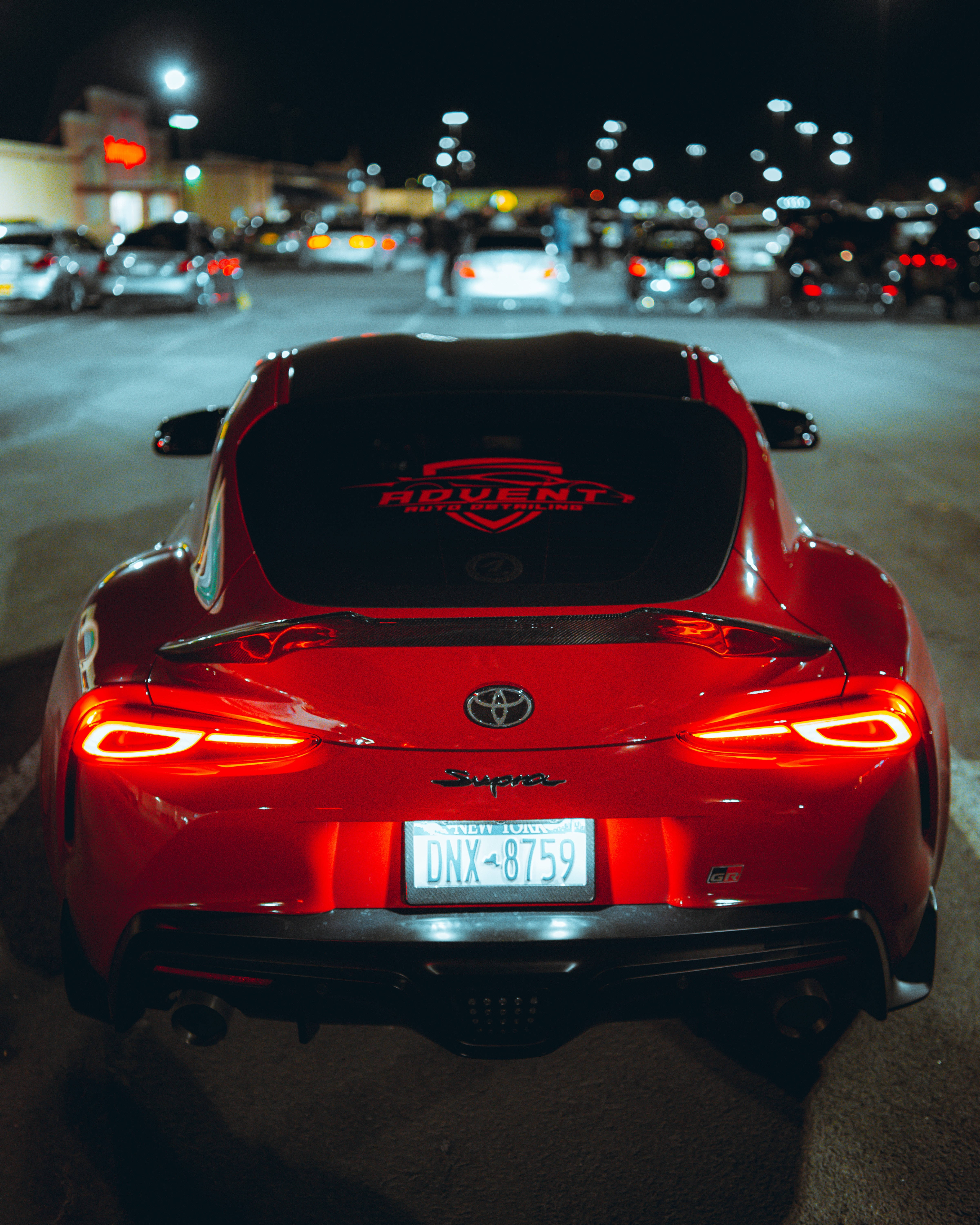 4K 《Toyota Supra》紅色車款搭配「Advent」 壁紙 — 一輛亮紅色豐田Supra跑車，尾燈發光，停泊在夜間城市停車場，背景燈光模糊。