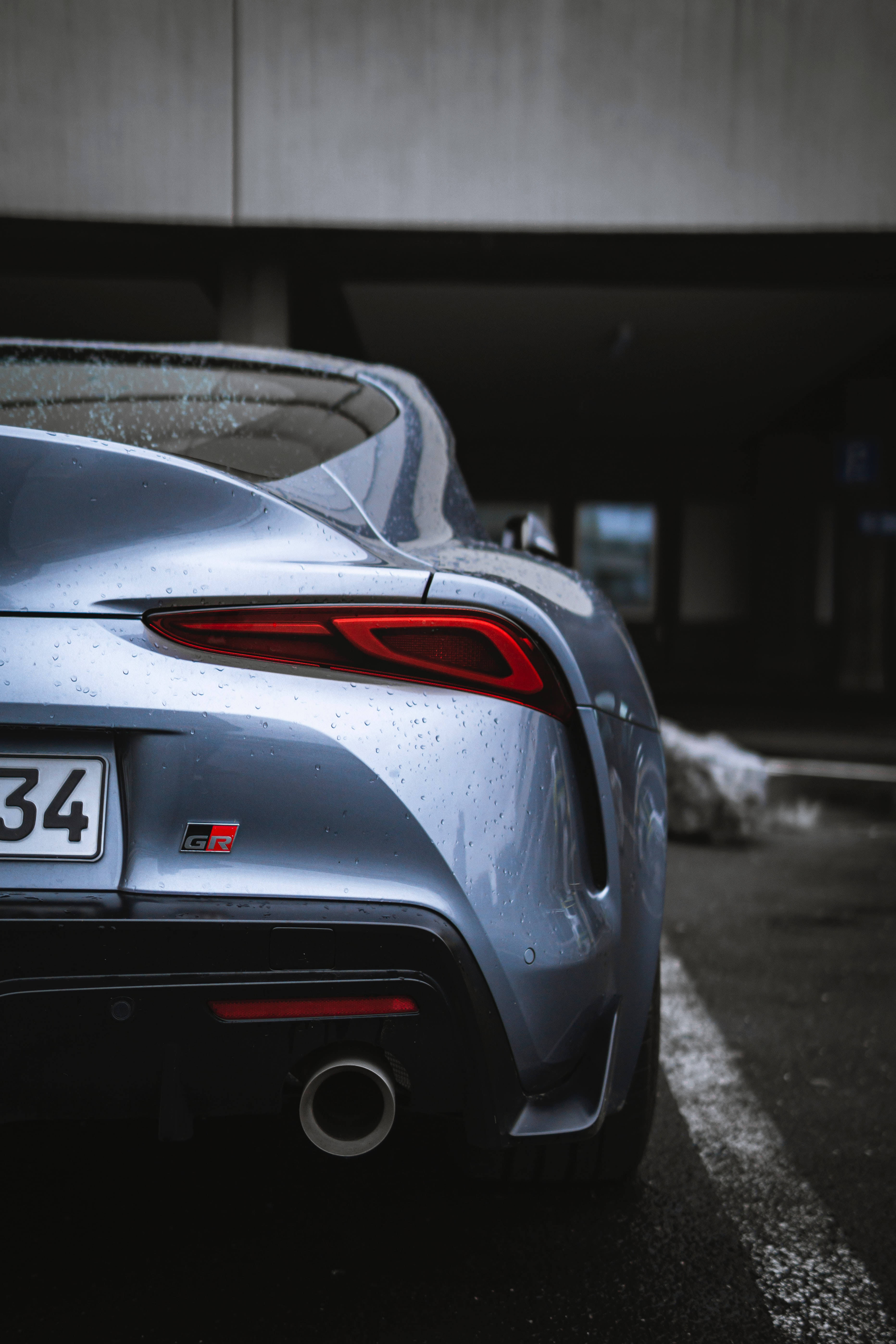 4K Toyota Supra GR灰色 壁紙 — 一架時尚的銀色跑車，紅色尾燈，停泊在混凝土天橋下，正值雨天。