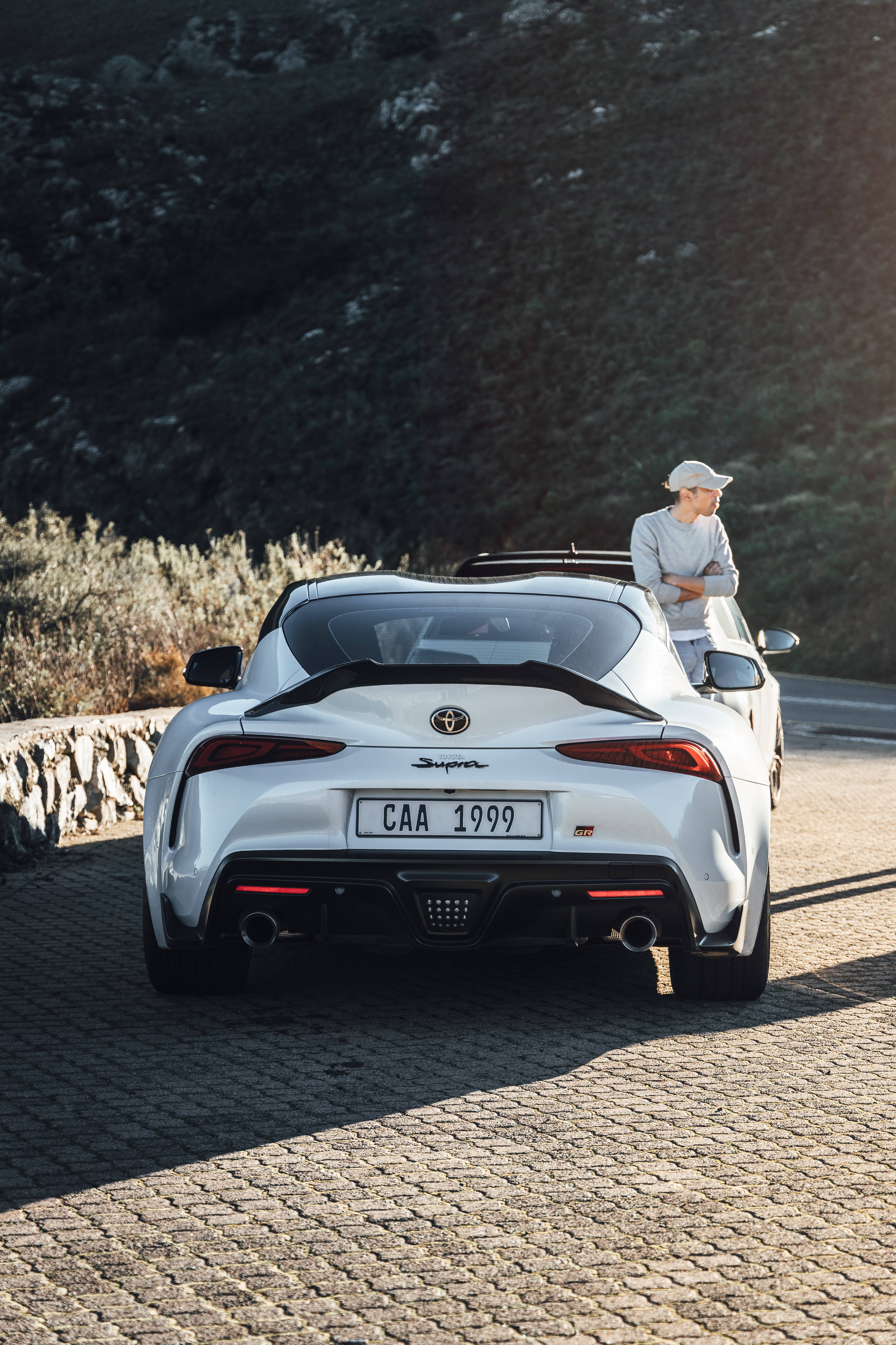 4K 《Toyota Supra》GR 白色車款 壁紙 — 一輛白色豐田Supra跑車停泊在陽光照耀的鵝卵石路上，一名戴帽子的男子靠在車上，背景是崎嶇的山丘。