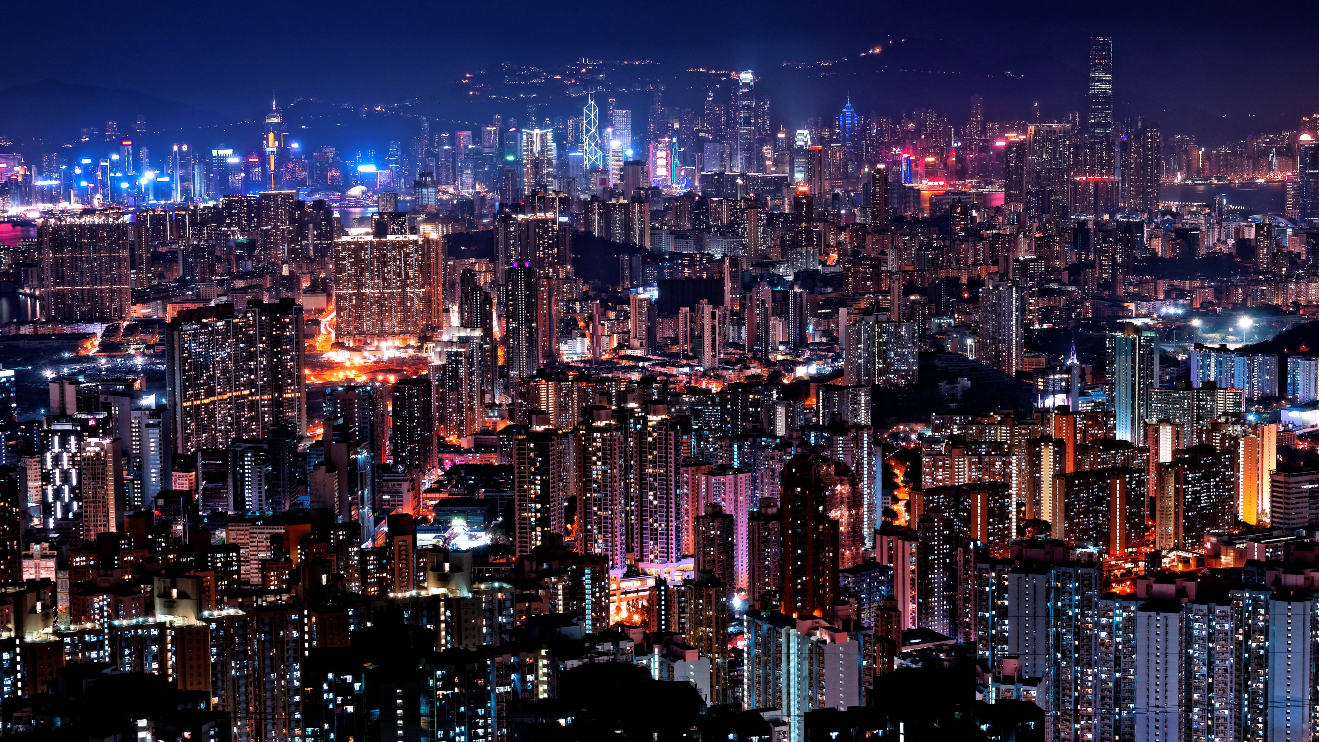 4K超高清香港城市明亮建築 壁紙 — 令人眼花繚亂的夜景城市景觀，高聳的摩天大樓和密集的住宅建築閃爍著鮮豔的霓虹燈光。