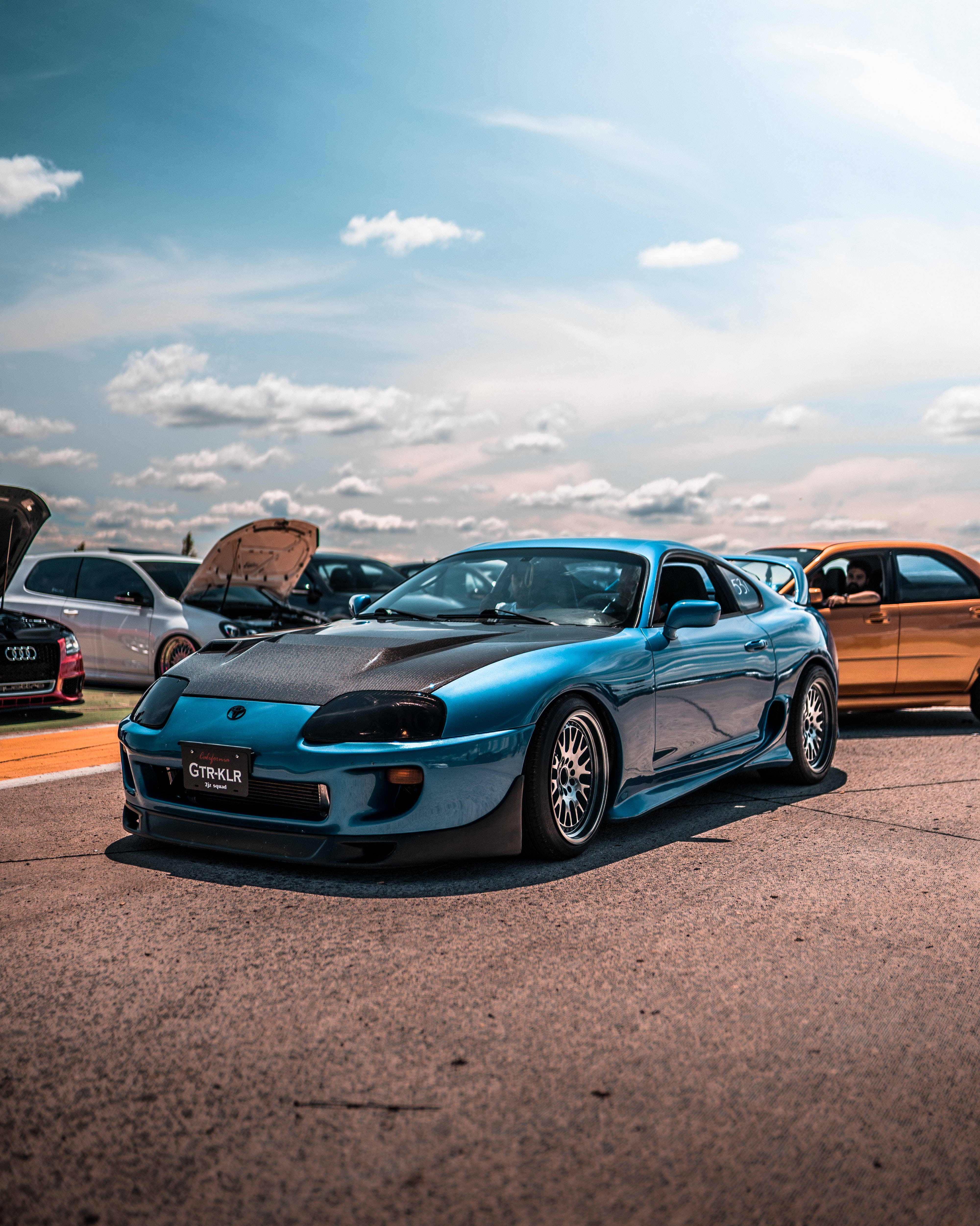 4K 丰田Supra 時尚跑車 壁紙 — 一輛鮮藍色改裝跑車，黑色引擎蓋，在明亮局部多雲的天空下，顯眼地停在瀝青場地上。