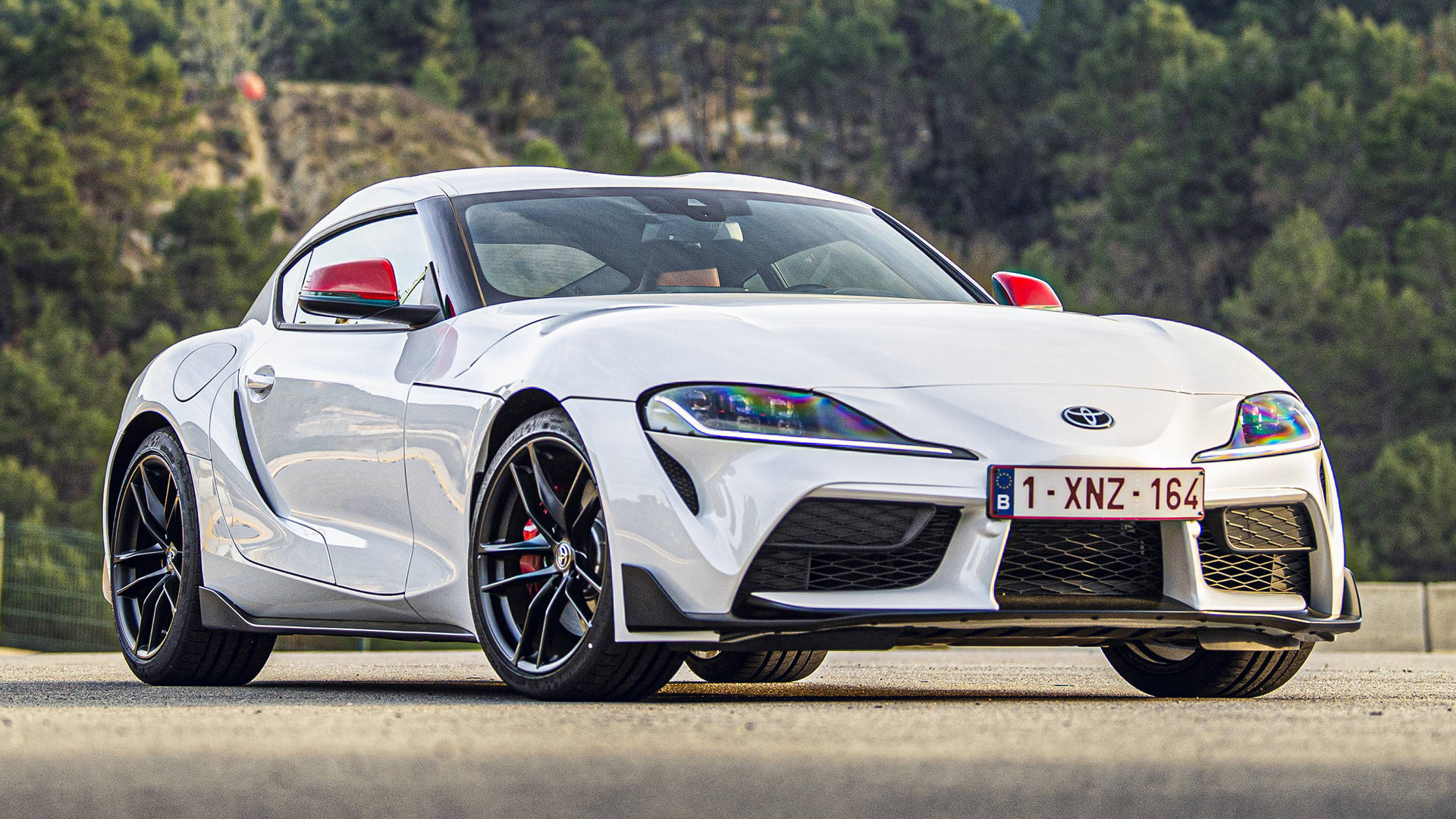 4K 丰田Supra 白色配紅色後視鏡 壁紙 — 一輛時尚的白色豐田Supra跑車，帶有紅色點綴，停泊在鋪砌的道路上，周圍綠樹成蔭。