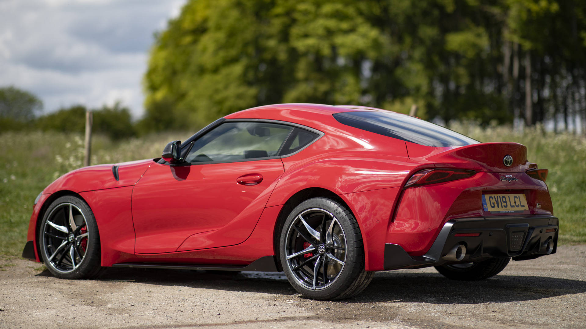 4K 丰田Supra 紅色停泊於樹林旁 壁紙 — 一輛鮮紅色的豐田Supra跑車停在碎石上，背景是茂密的綠樹，天空多雲。