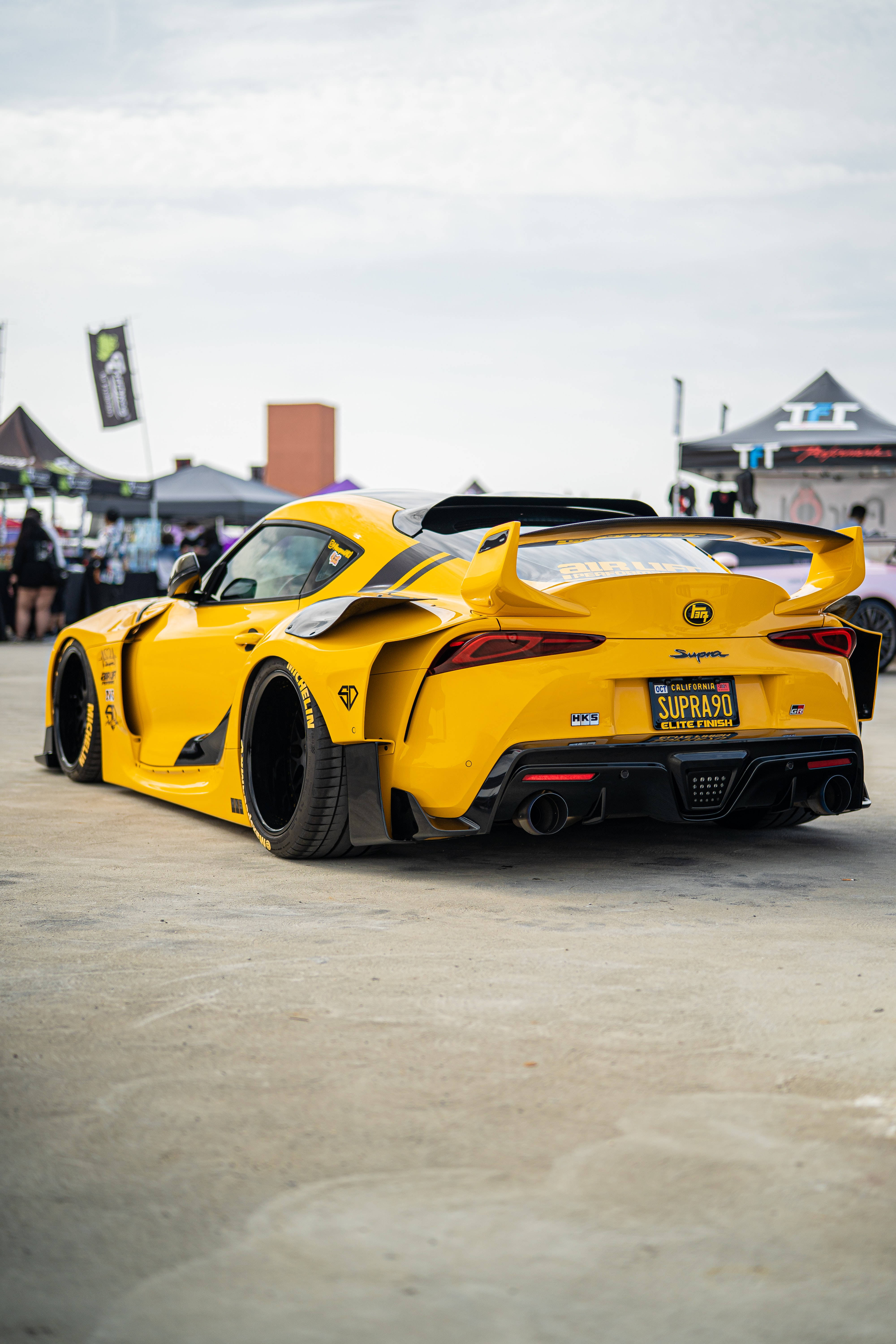 4K 丰田Supra 黃色車款 壁紙 — 一架鮮黃色改裝豐田Supra跑車，帶有大型尾翼，停泊在戶外車展，天空多雲。