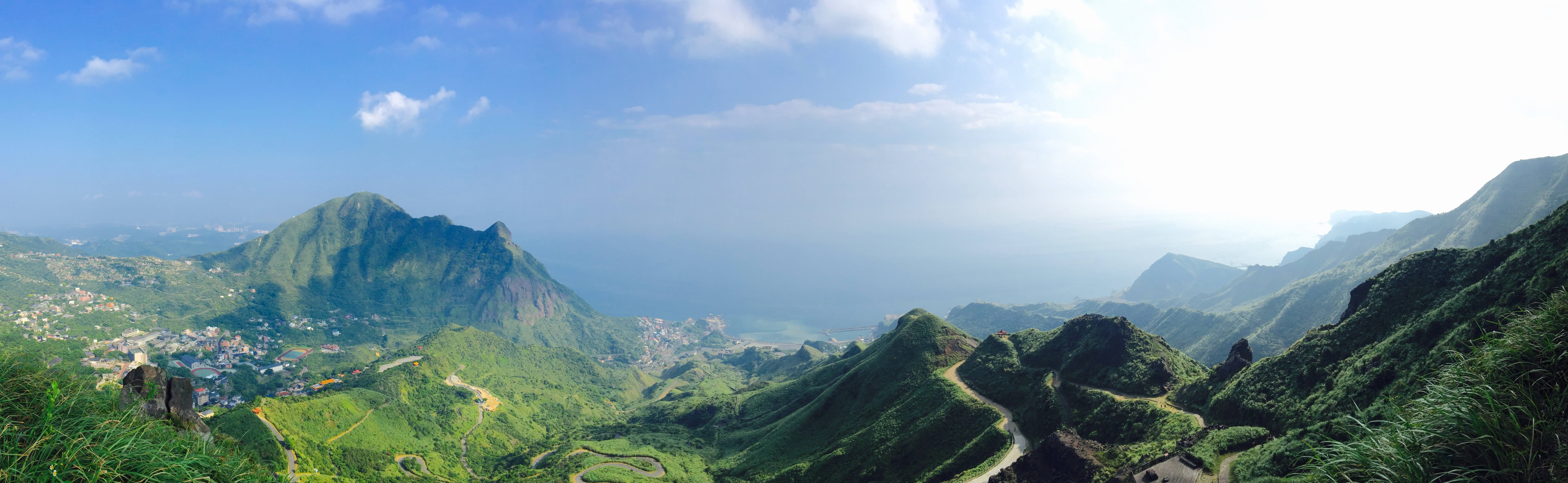 4K 双螢幕青草地山景 壁紙 — 連綿起伏的翠綠山脈、蜿蜒的道路和沿海村莊的全景，在明亮的藍天下點綴著幾朵白雲。