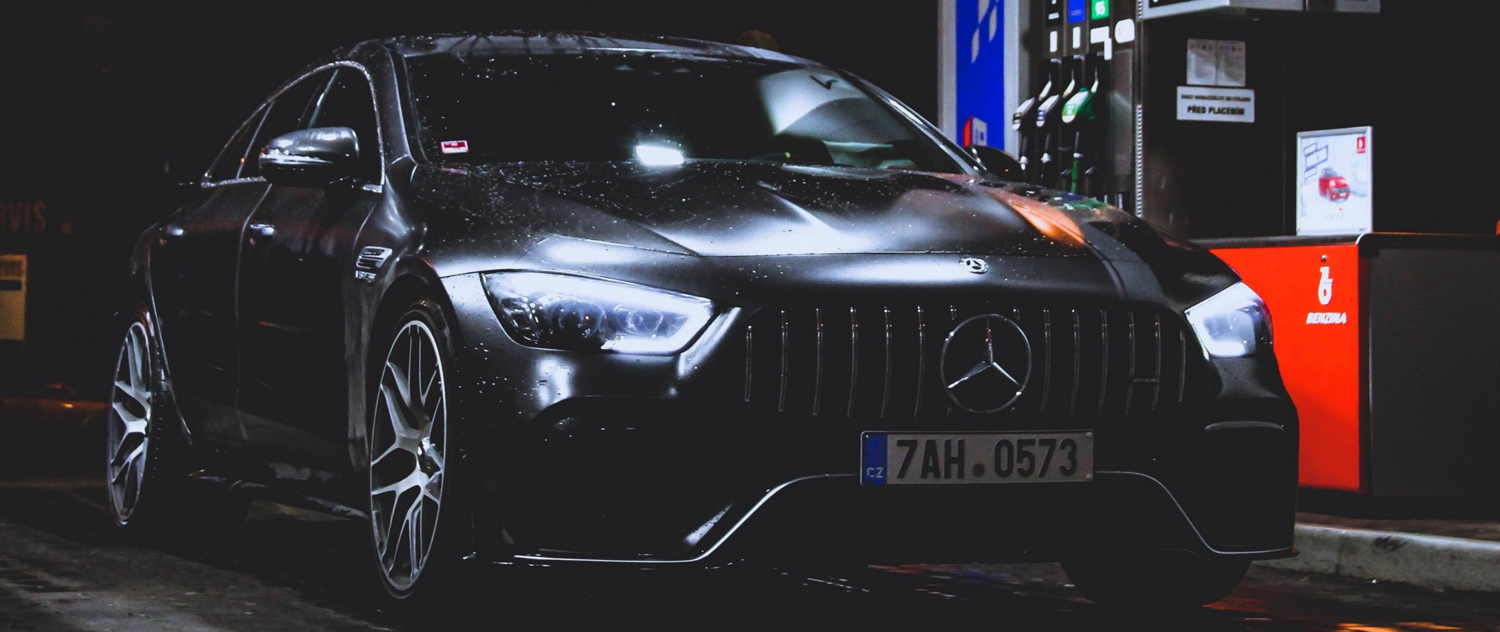 4K 夜間街頭黑奔馳 壁紙 — 一輛時尚的黑色Mercedes-AMG在夜間停放在加油站，光滑表面上有雨滴，車頭燈發光。