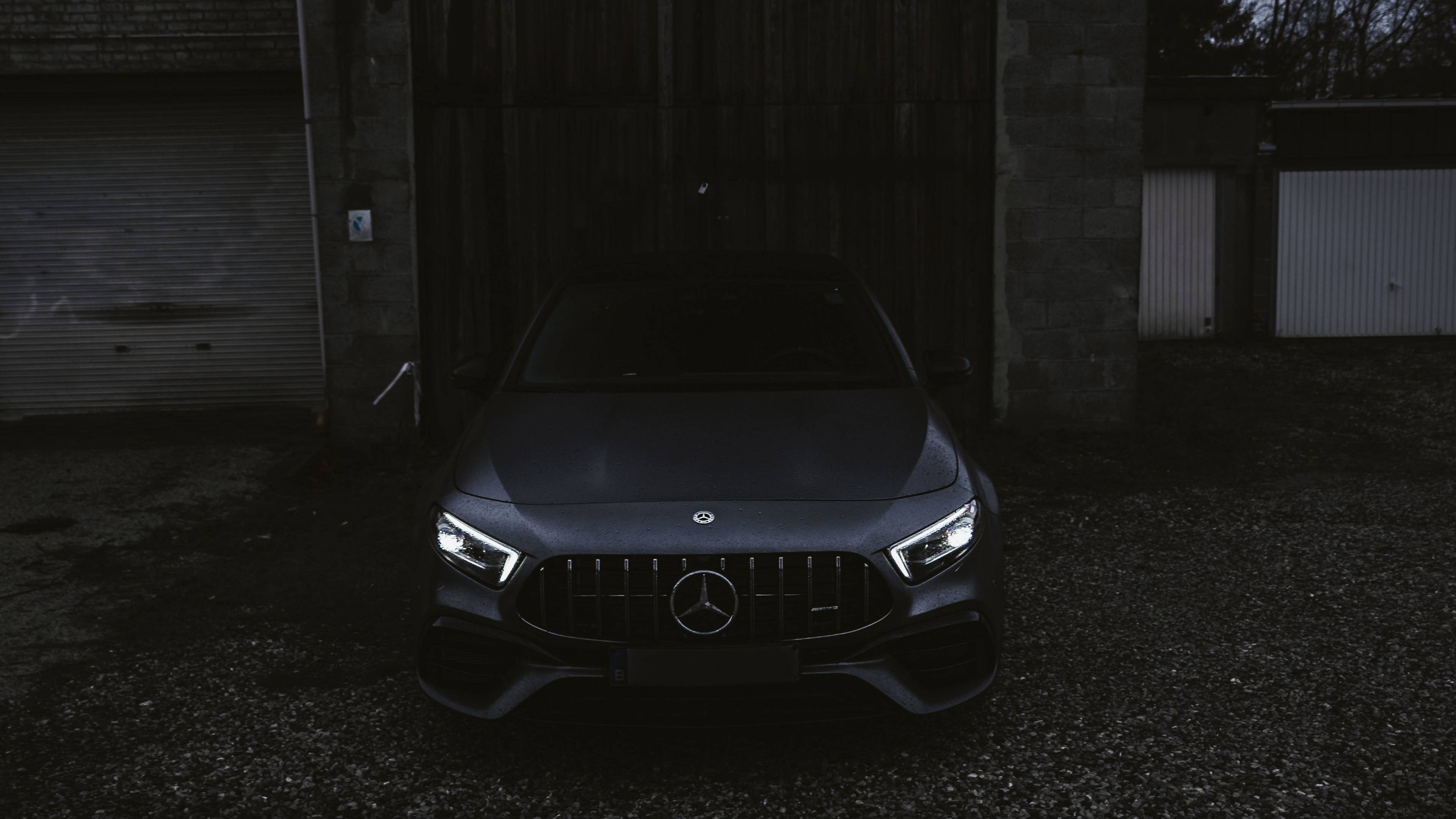 4K 奔馳 A 級 壁紙 — 一輛時尚的深灰色Mercedes-AMG跑車，車頭燈發光，夜間停放在風化的工業車庫前。
