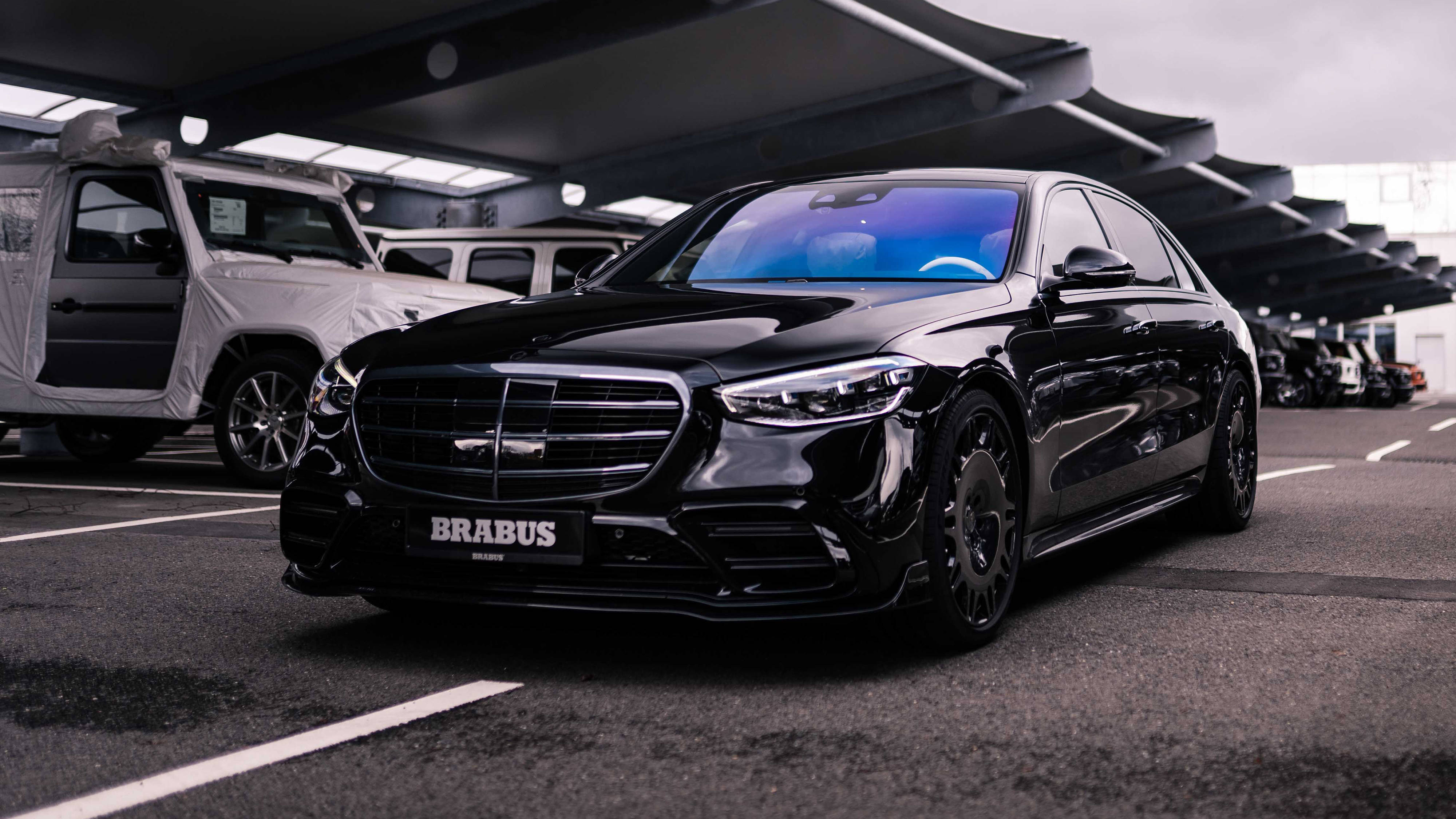 4K 奔馳停泊於停車場 壁紙 — 一輛光澤黑色Brabus Mercedes轎車在陰天下的現代有蓋停車場中閃耀。