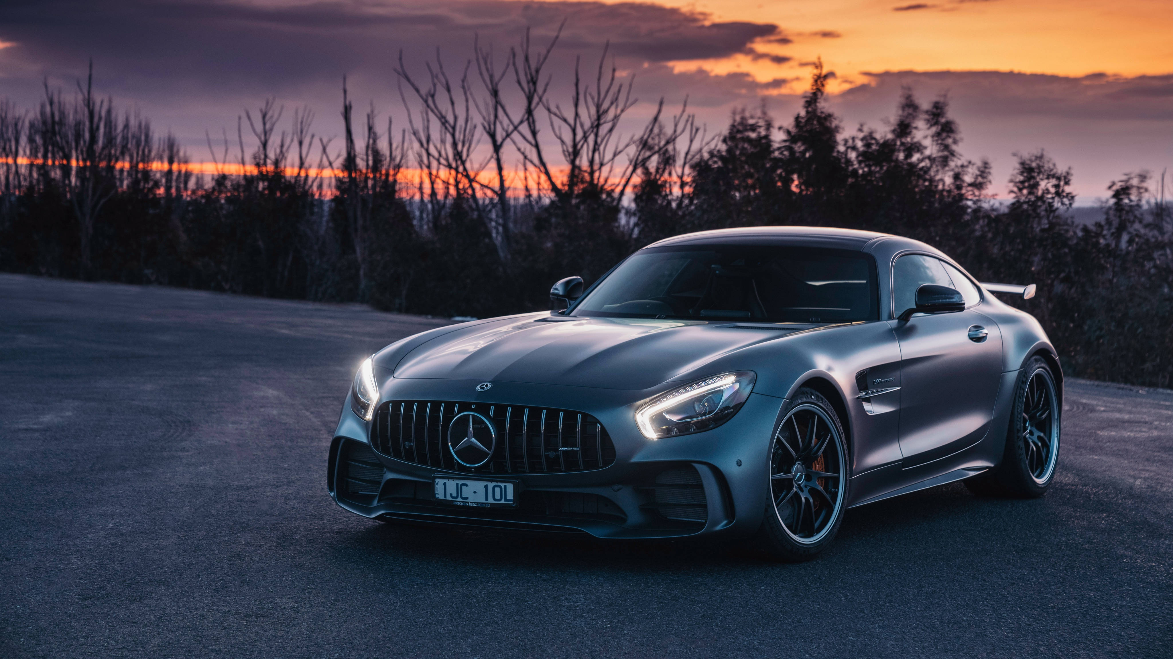 4K 奔馳樹林背景 壁紙 — 一架時尚的銀色 Mercedes-AMG GT 停泊在日落時分的道路上，背景有光禿的樹木和充滿活力的橙色天空。