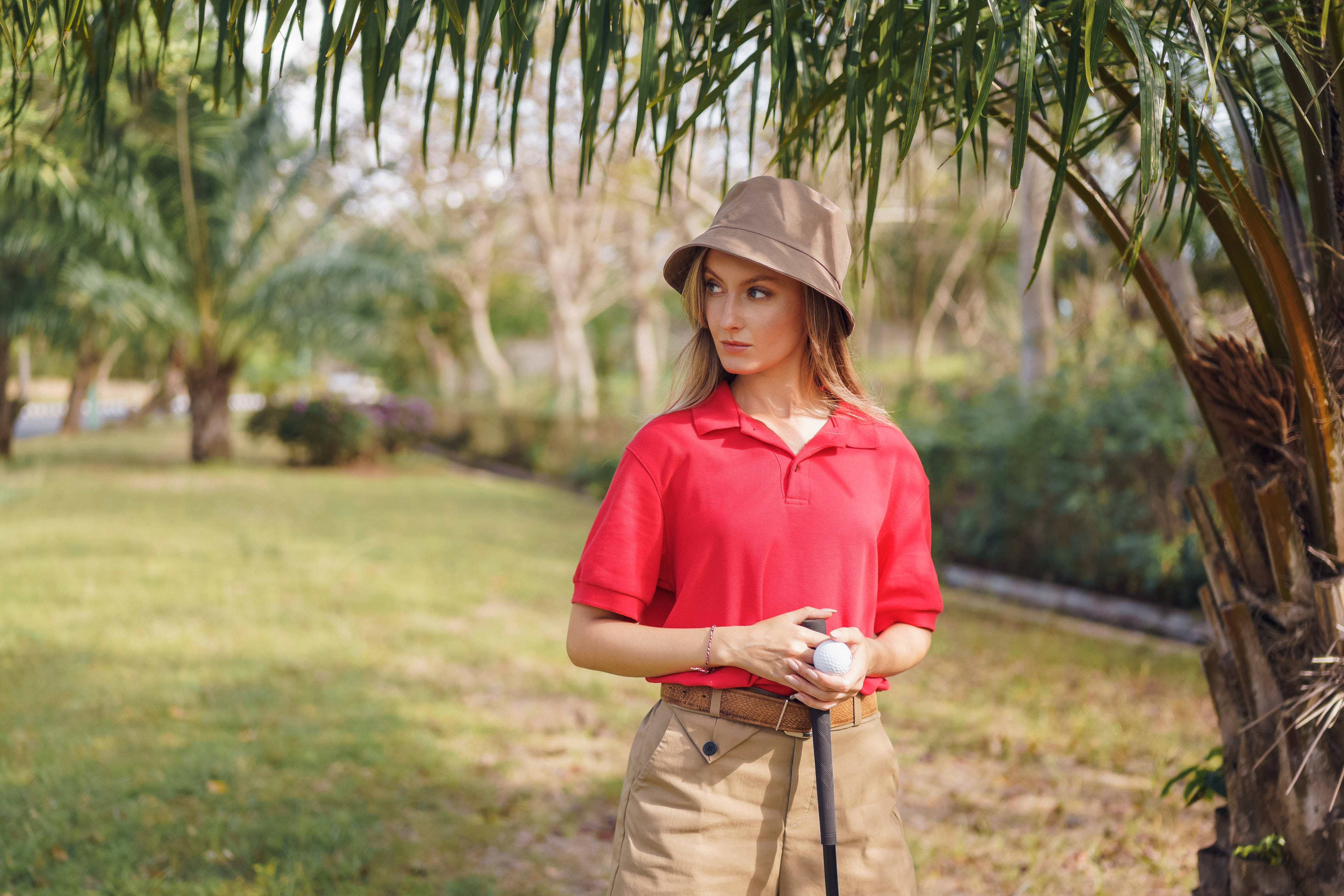 4K 女士手持高爾夫球 壁紙 — 一位身穿紅色Polo衫和米色帽子的女士，在陽光明媚的高爾夫球場上，棕櫚葉下拿著高爾夫球桿和球。