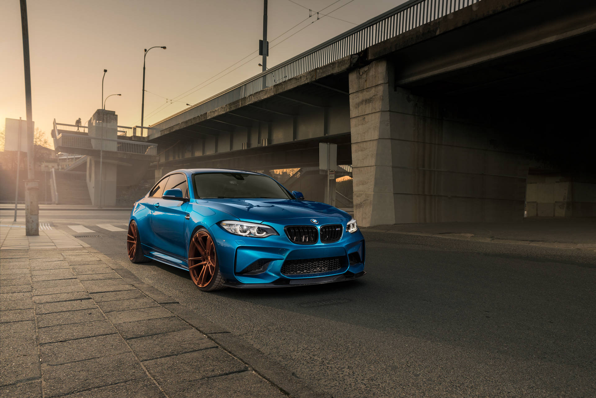 日落時分，一輛藍色BMW M2雙門跑車配備銅色輪圈，停泊在高架橋下。 — 4K BMW 壁紙 壁紙