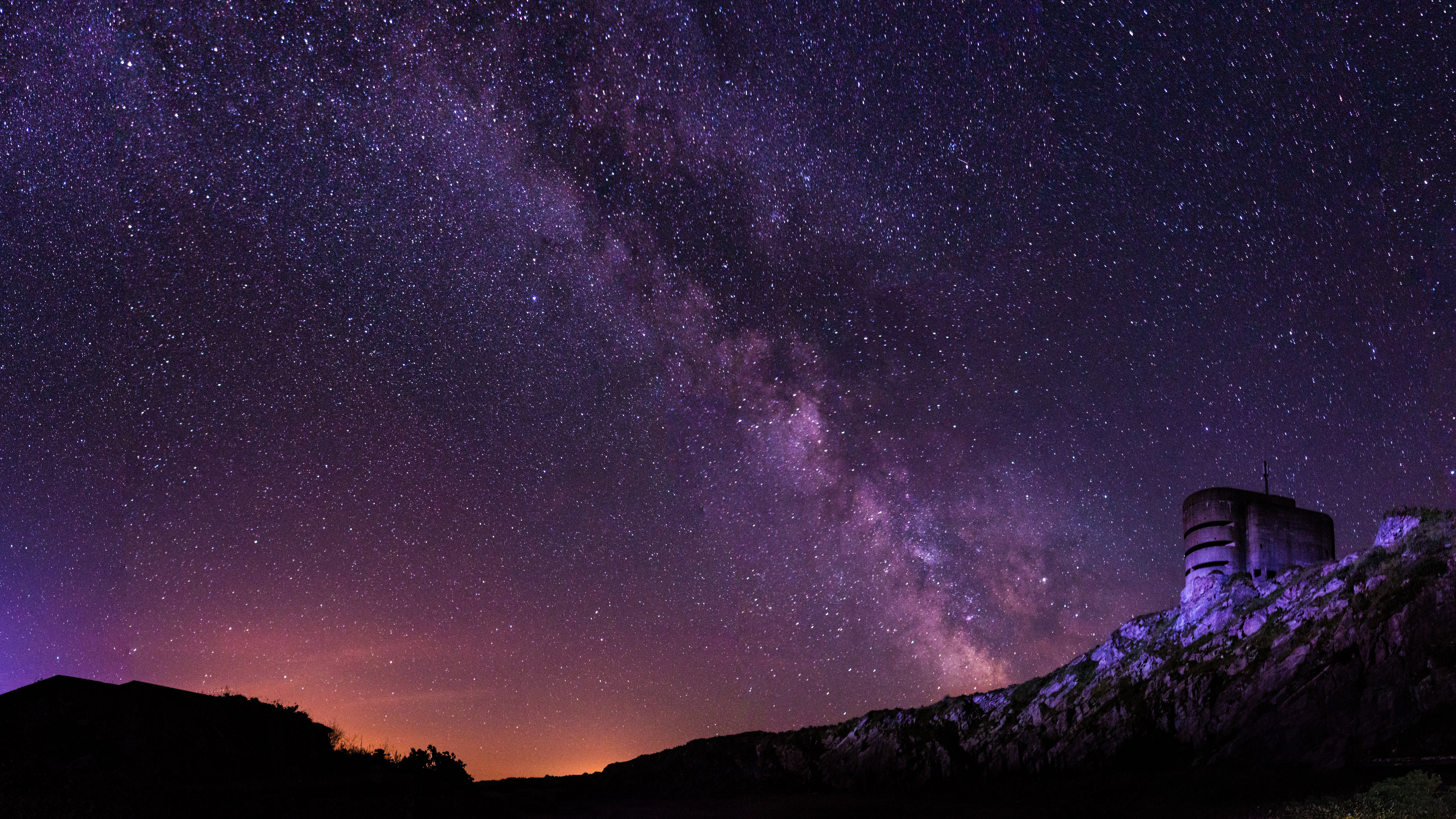 4K 星空桌面背景 壁紙 — 壯麗的紫色和橙色星空拱起在崎嶇山頂堡壘的輪廓之上。