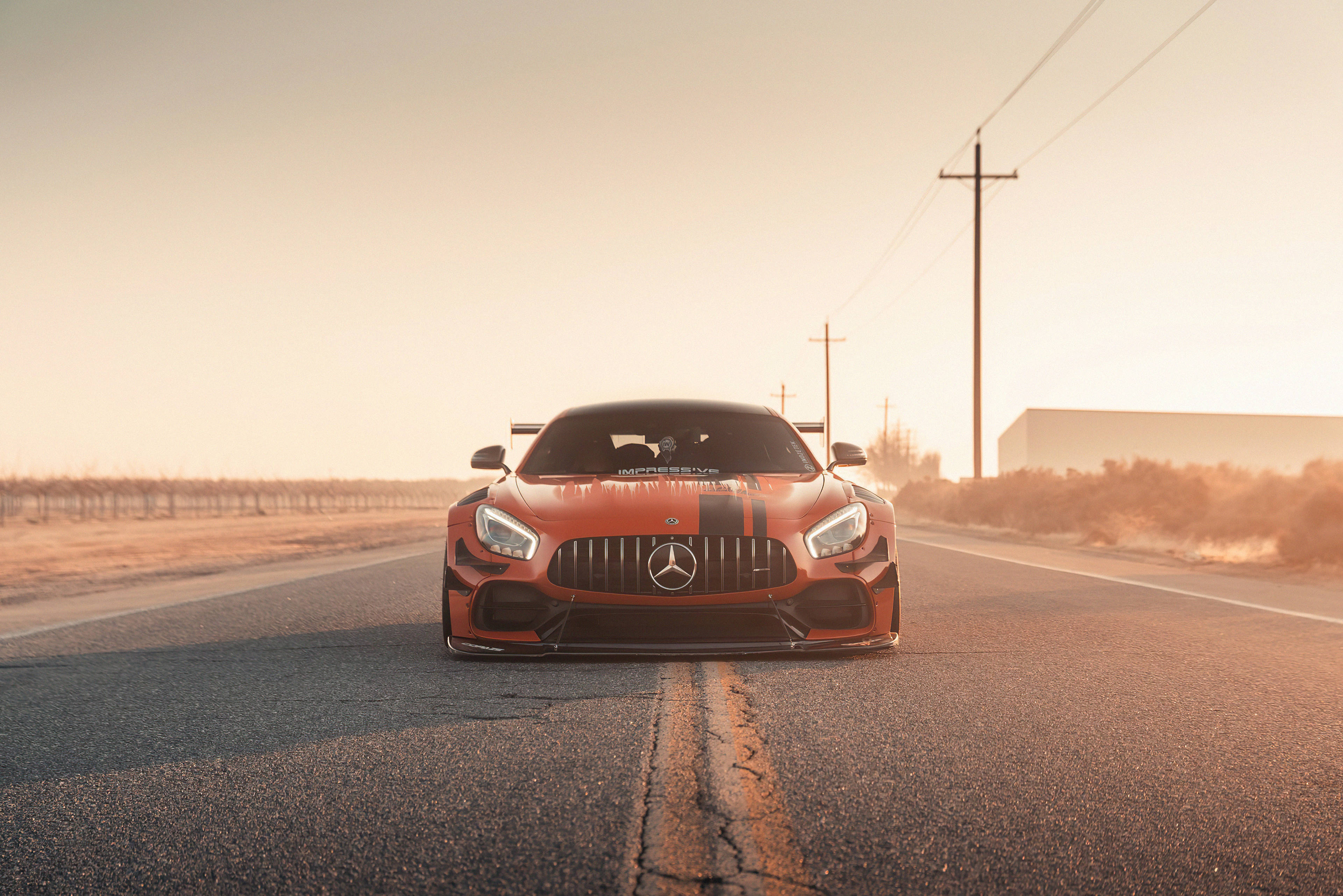 4K 梅賽德斯-奔馳行駛於道路 壁紙 — 一輛火橙色的Mercedes-AMG GT R在黃金時段面向前方，行駛在裂開的瀝青路上，背景有電線和乾燥的田野。