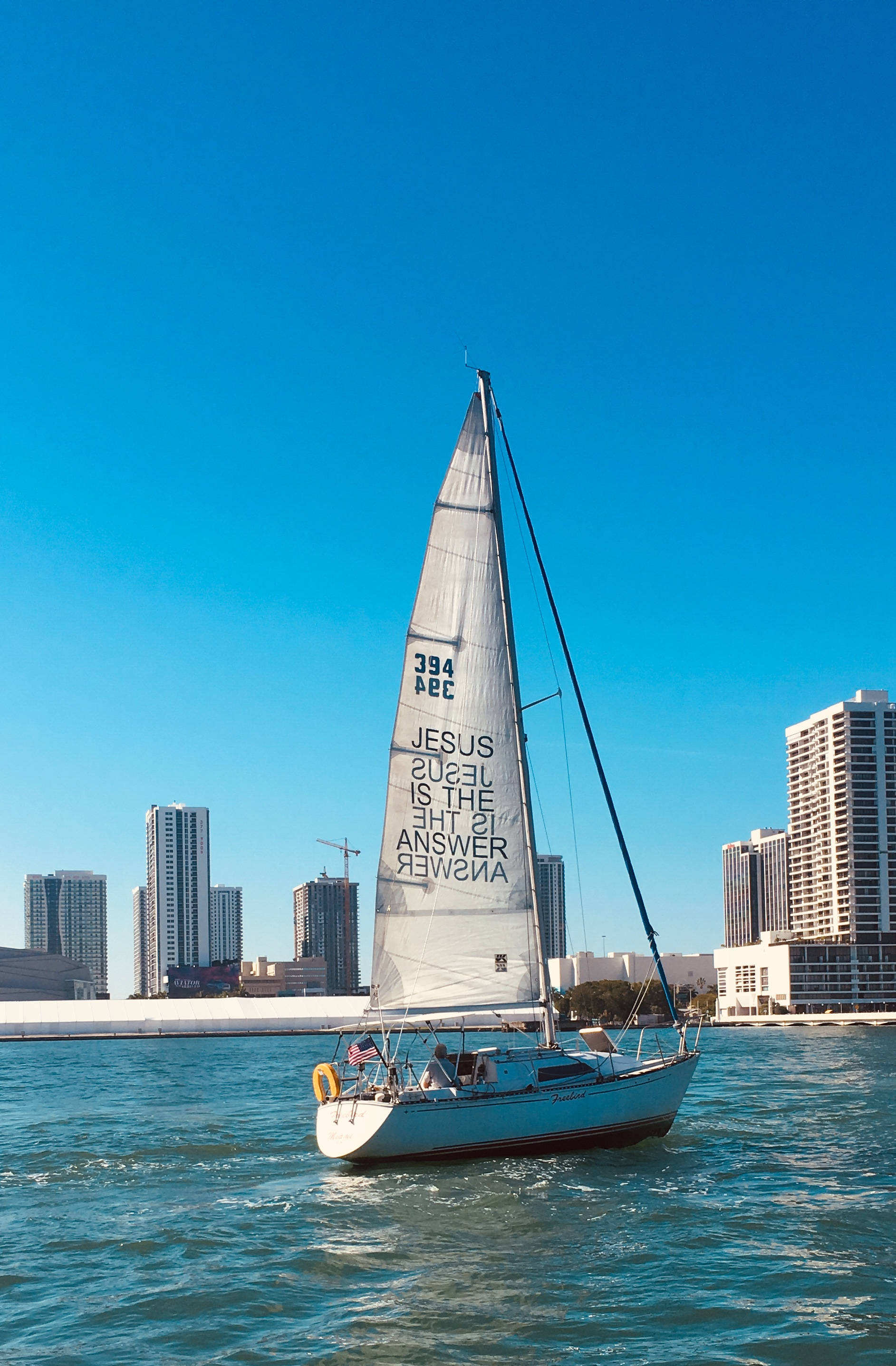 4K 海灘帆船 壁紙 — 一艘白色帆船帶有粗體文字，在碧綠的水面上平靜航行，背景是現代城市摩天大樓，天空清澈湛藍。