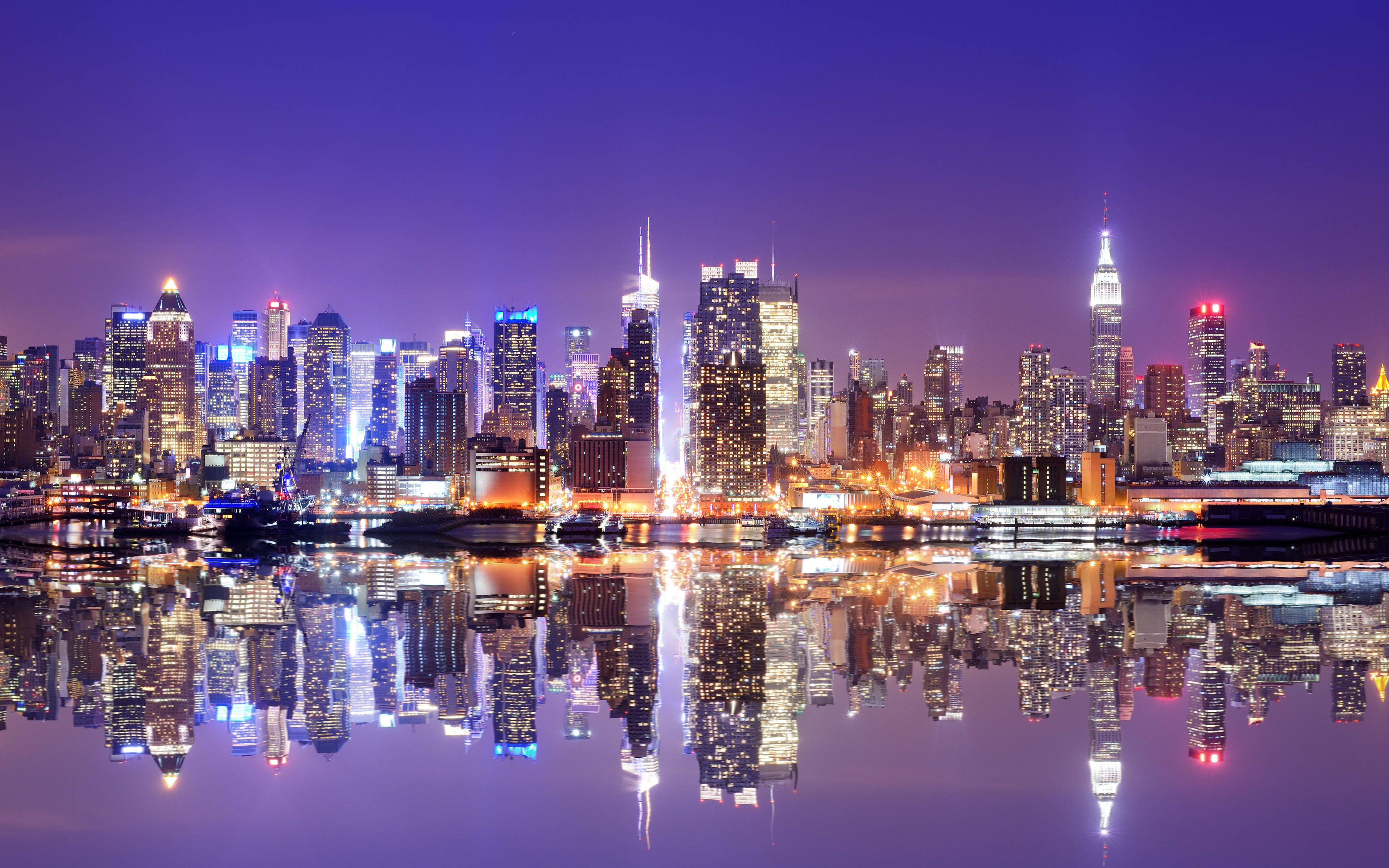4K 紐約曼哈頓倒影 壁紙 — 紐約令人目眩的夜景，摩天大樓在深紫色天空下閃耀，完美倒映在下方平靜的水面上。