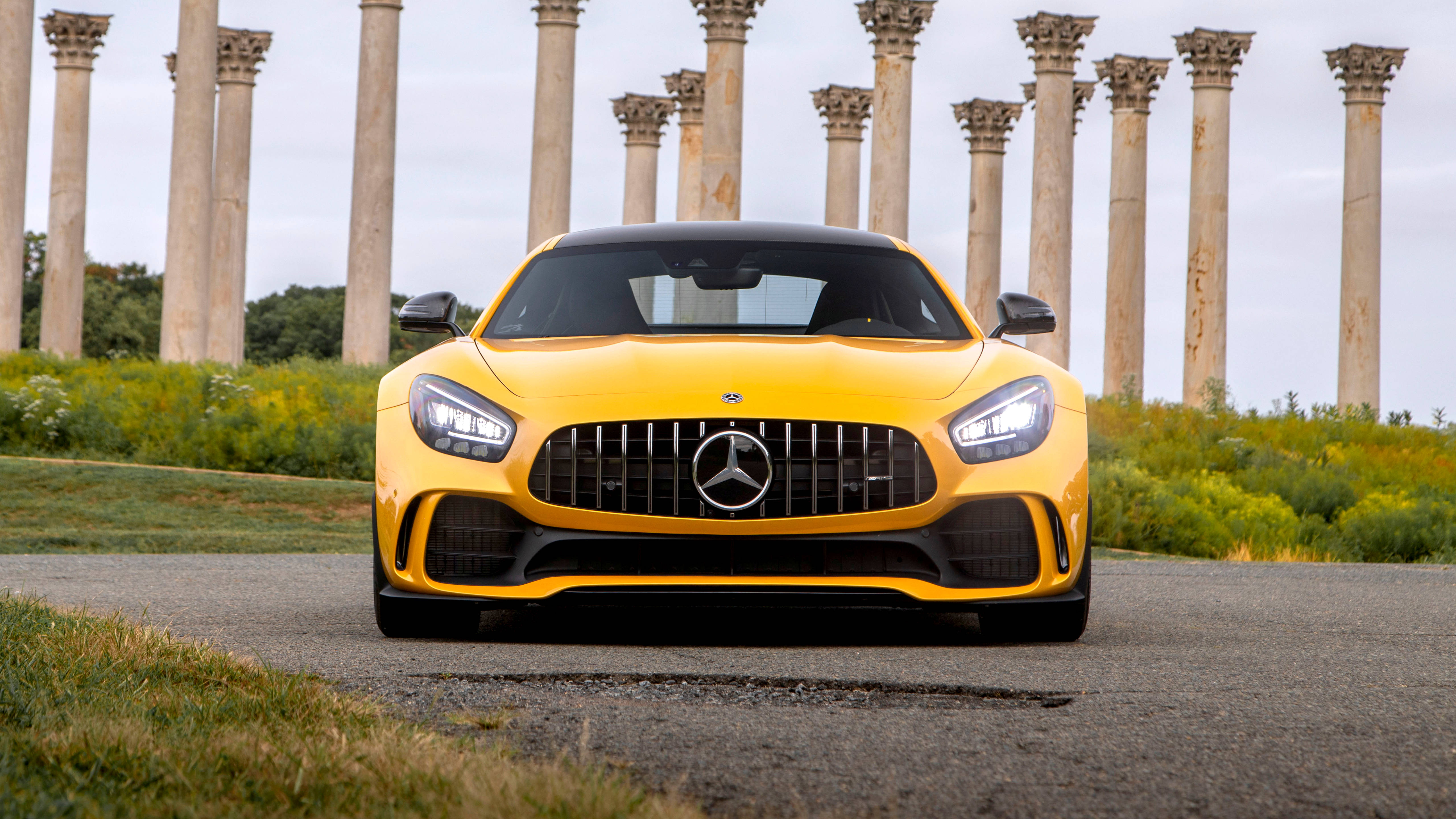4K 黃色奔馳配混凝土柱 壁紙 — 一輛鮮黃色的 Mercedes-AMG GT 跑車在陰天之下，面向前方，停泊在由古老石柱環繞的鋪砌小徑上。