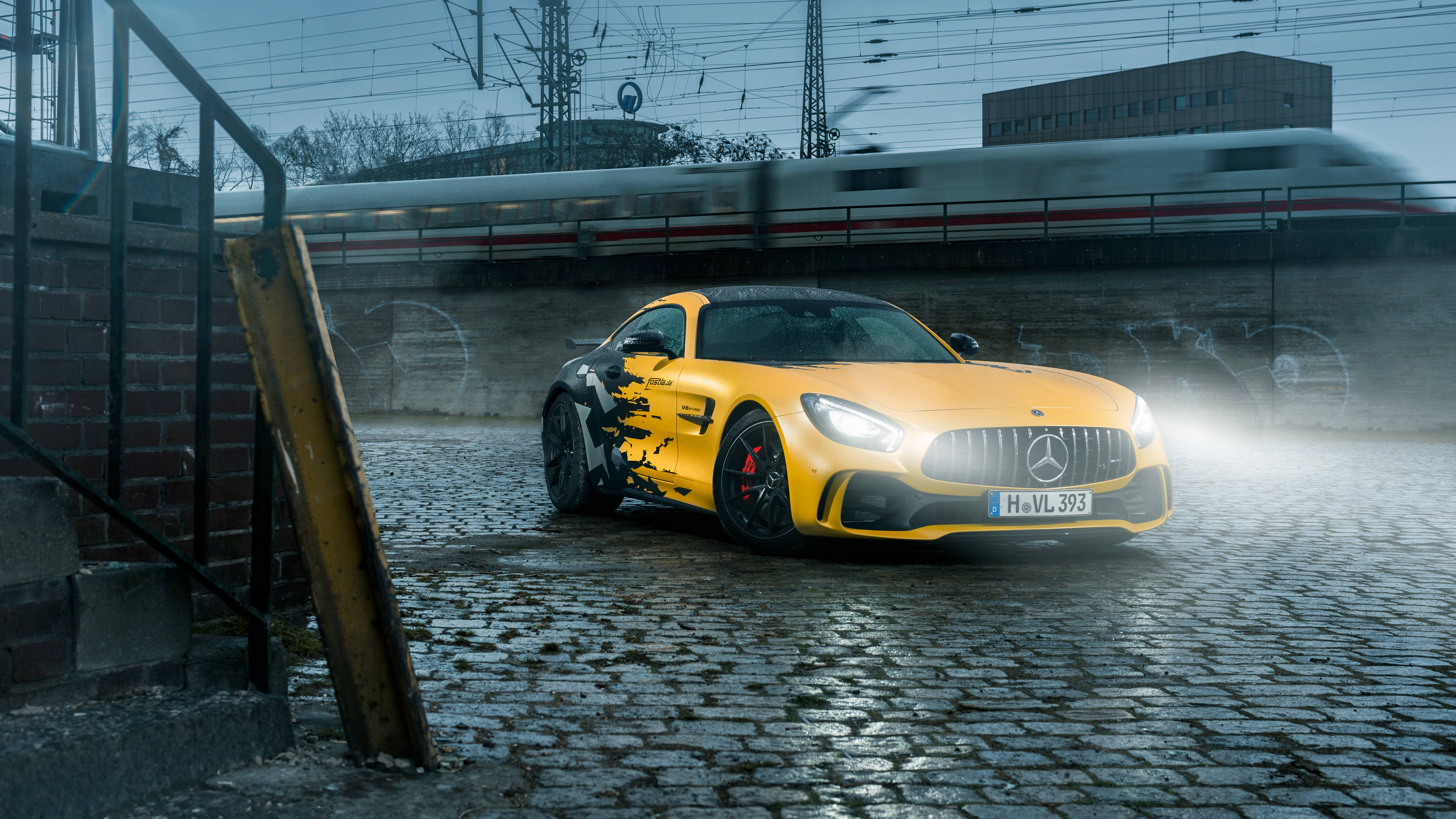 4K 黃黑配色奔馳 壁紙 — 一輛鮮黃色的 Mercedes-AMG GT 跑車帶有黑色火焰貼紙，停在濕漉漉的鵝卵石上，一列模糊的高速列車在陰天從上方駛過。
