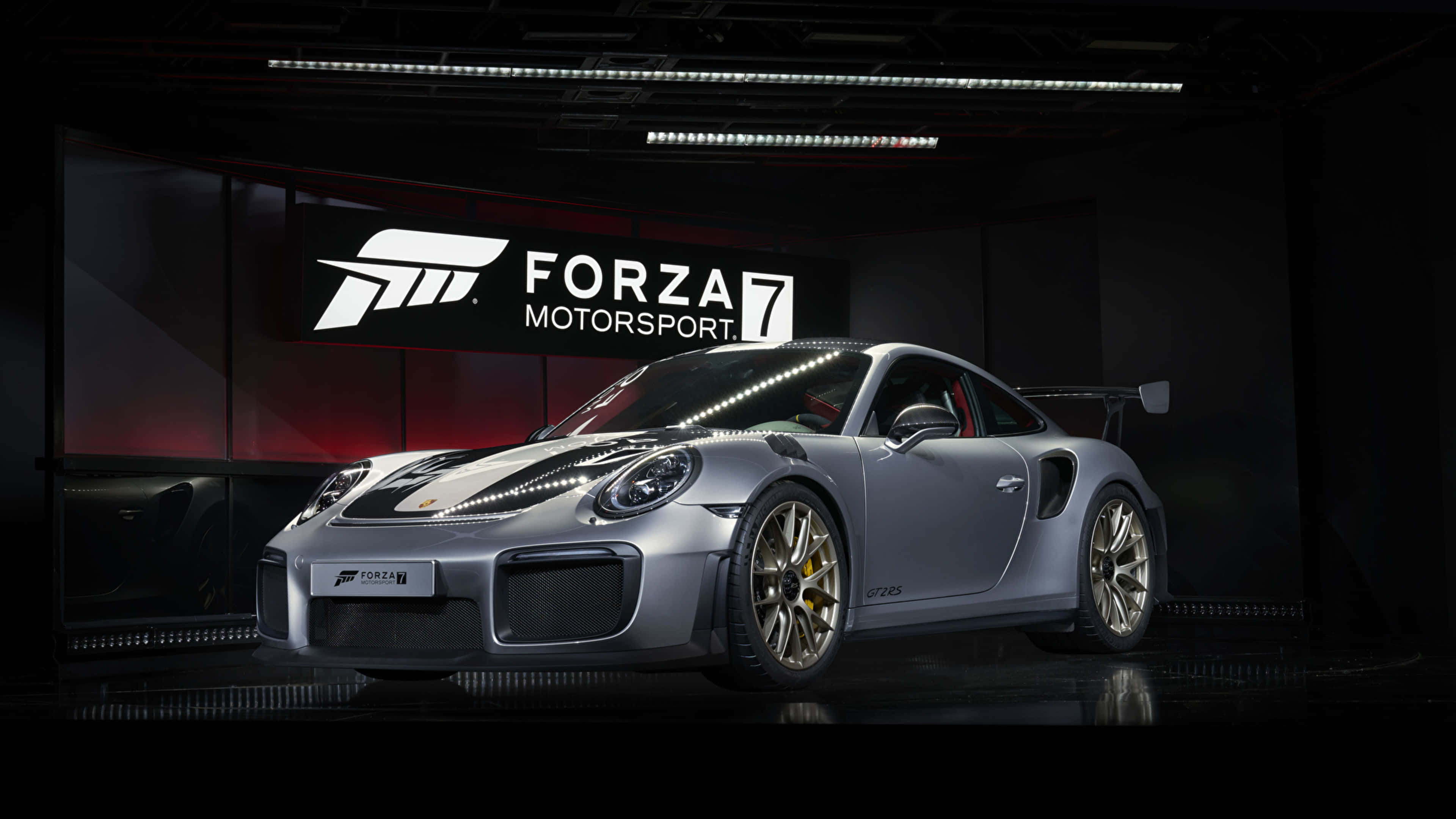 以 4K《極限競速7》衝向終點線 壁紙 — 一輛時尚的銀色保時捷911 GT2 RS配備金色輪圈，在黑暗的Forza Motorsport 7品牌車庫中於工作室燈光下展示。