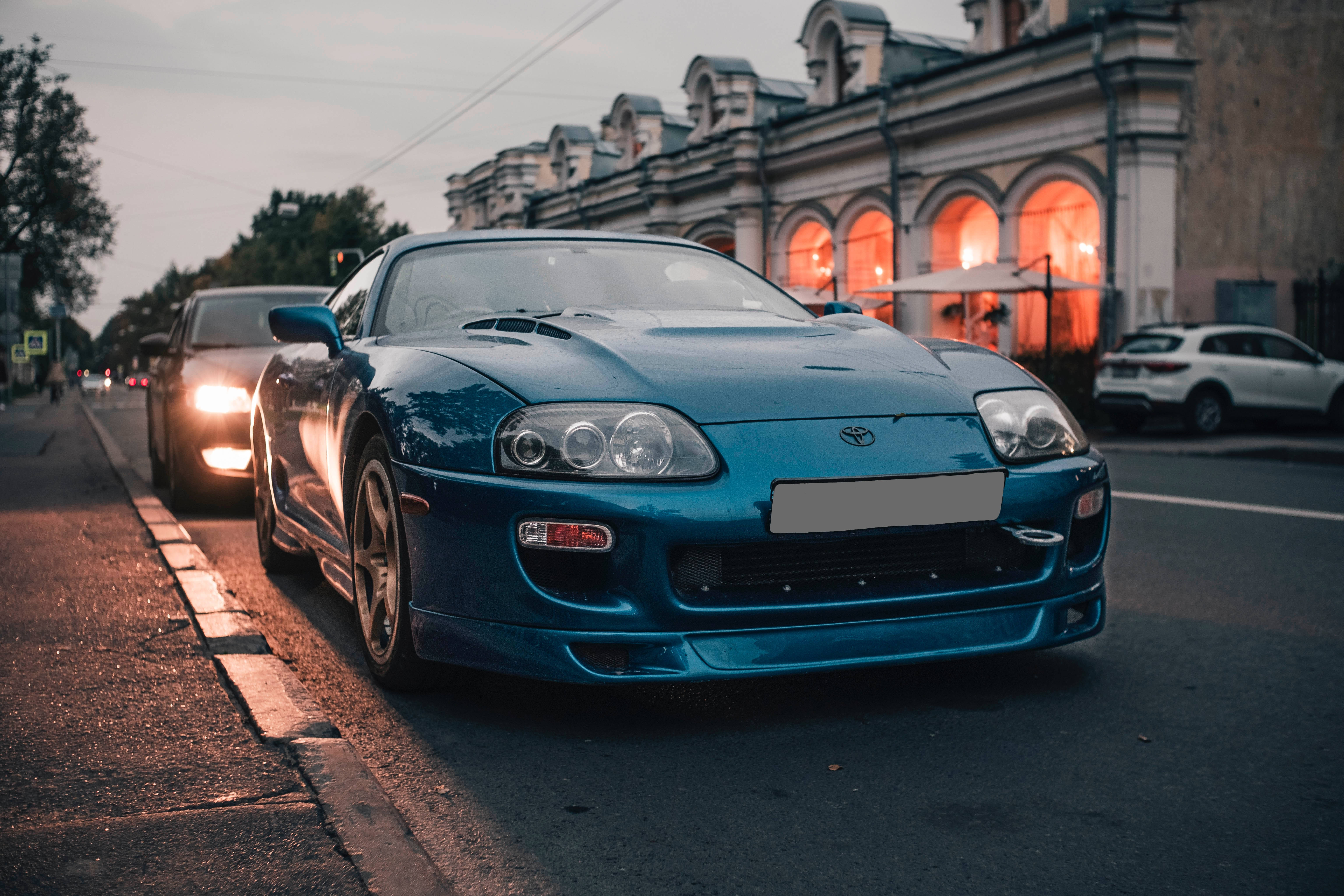 4K天藍色Toyota Supra 壁紙 — 一架時尚的藍色跑車停泊在黃昏的城市街道上，背後有溫暖發光的窗戶。