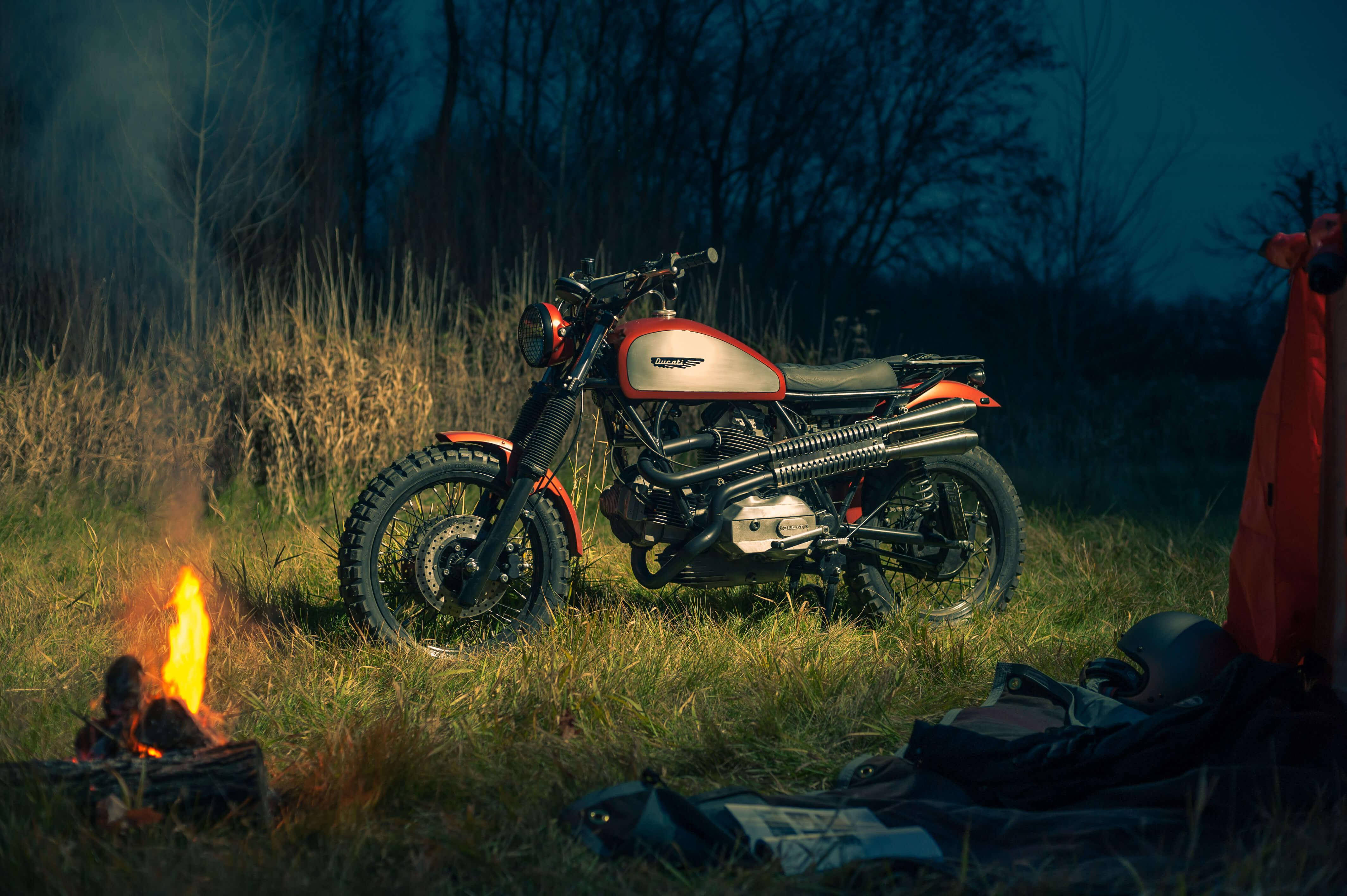 4K杜卡迪Scrambler機車 壁紙 — 一輛紅銀色電單車停泊在黃昏時分的草地上，旁邊有營火，附近有一個帳篷。