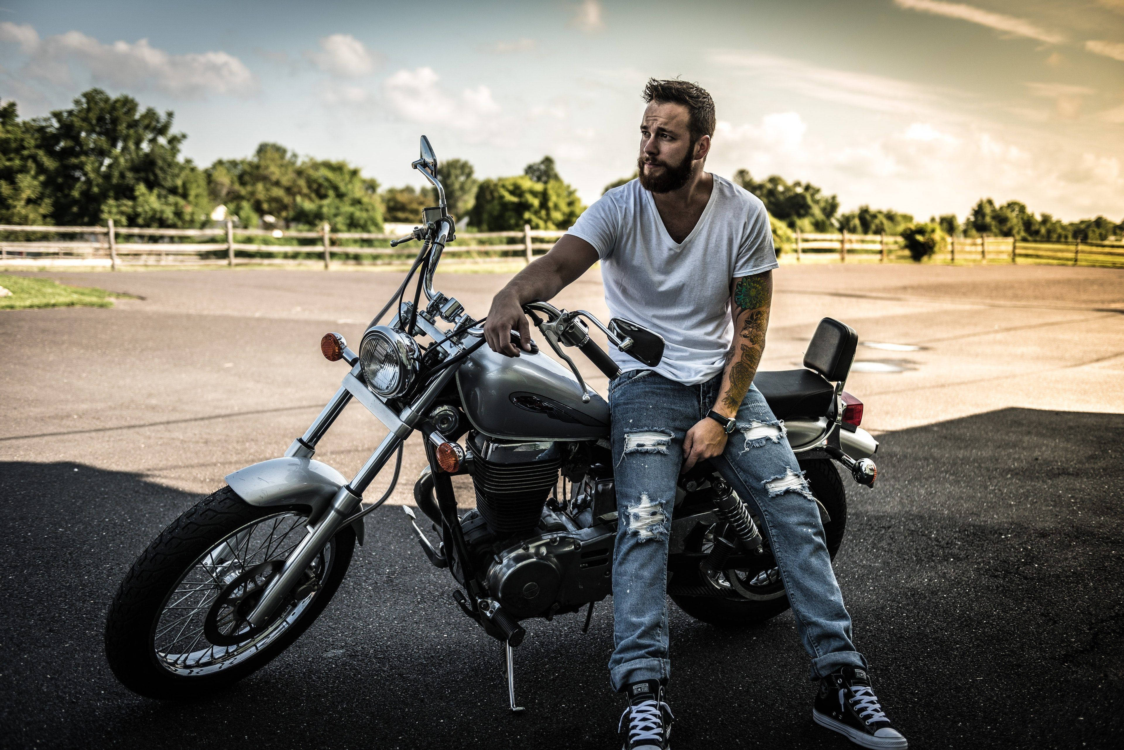 4K自行車與帥氣騎士 壁紙 — 一位留鬍鬚、有紋身的男子自信地坐在一輛停在瀝青路面上的銀色巡航電單車上，天空局部多雲。