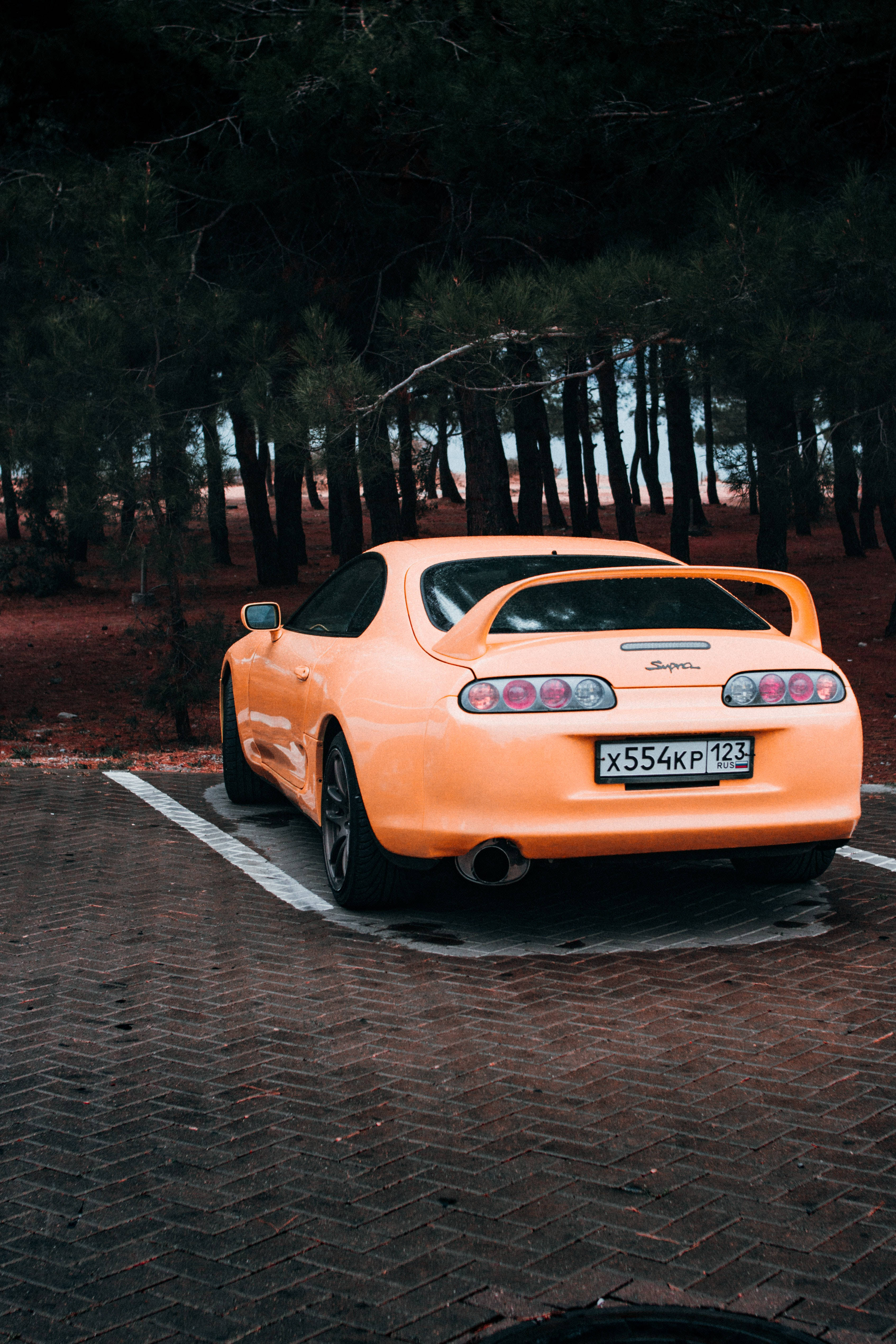 4K黃色Toyota Supra 壁紙 — 一架充滿活力的橙色跑車停泊在磚砌車道上，周圍是森林中的高大松樹。