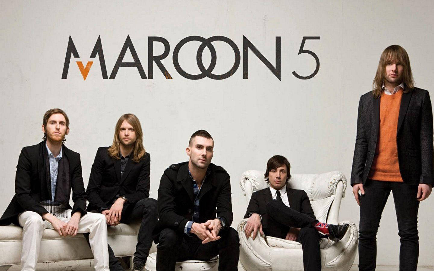馬龍5沙發椅擺拍 壁紙 — Maroon 5 樂隊成員自信地穿著黑色西裝和橙色點綴，在極簡白色牆壁前與他們的標誌合影。