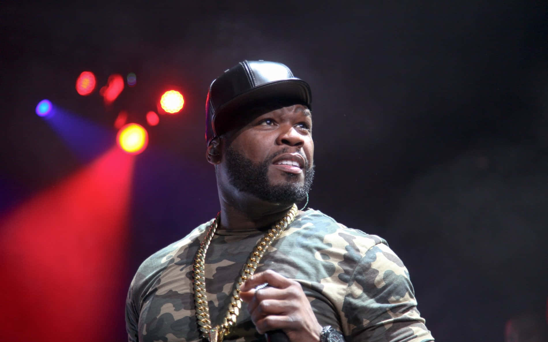 50 Cent 領銜戴「黑桃A」鑽石項鍊 壁紙 — 饒舌歌手50 Cent在舞台上表演，身穿迷彩襯衫和金鏈，在彩色舞台燈光下。