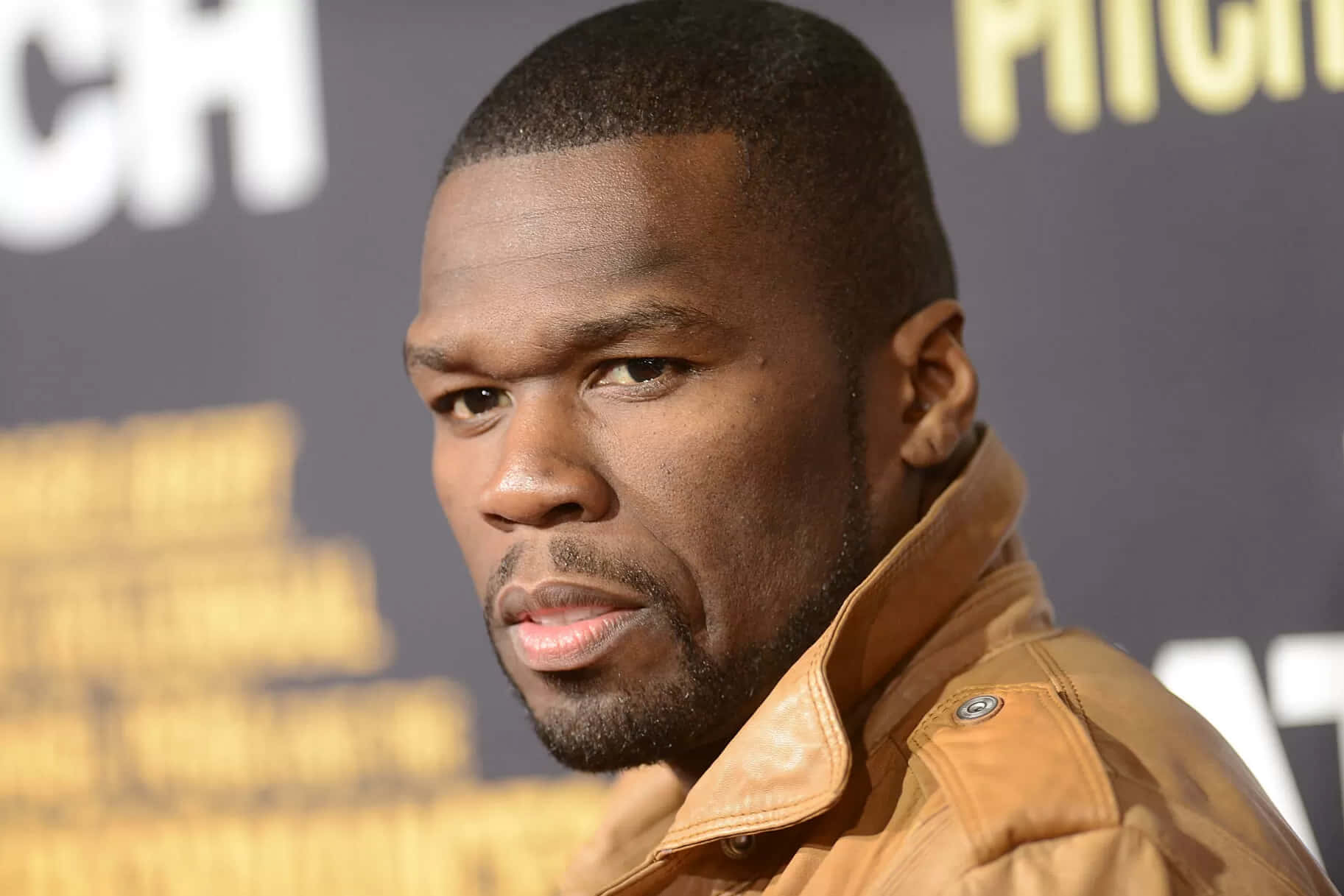 50 Cent 身穿棕褐色皮褸出席活動，背景為模糊文字。