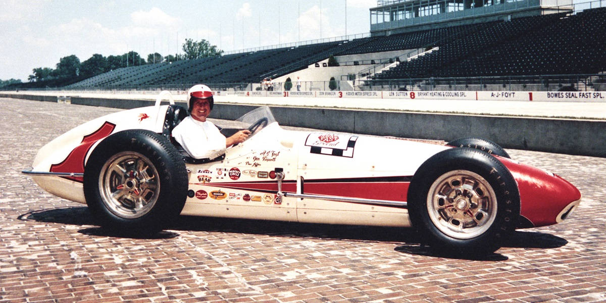 印第安納波利斯500 賽車手 AJ Foyt 壁紙 — 一輛復古白色和紅色賽車，司機在明亮的日光下在磚砌賽道上微笑。