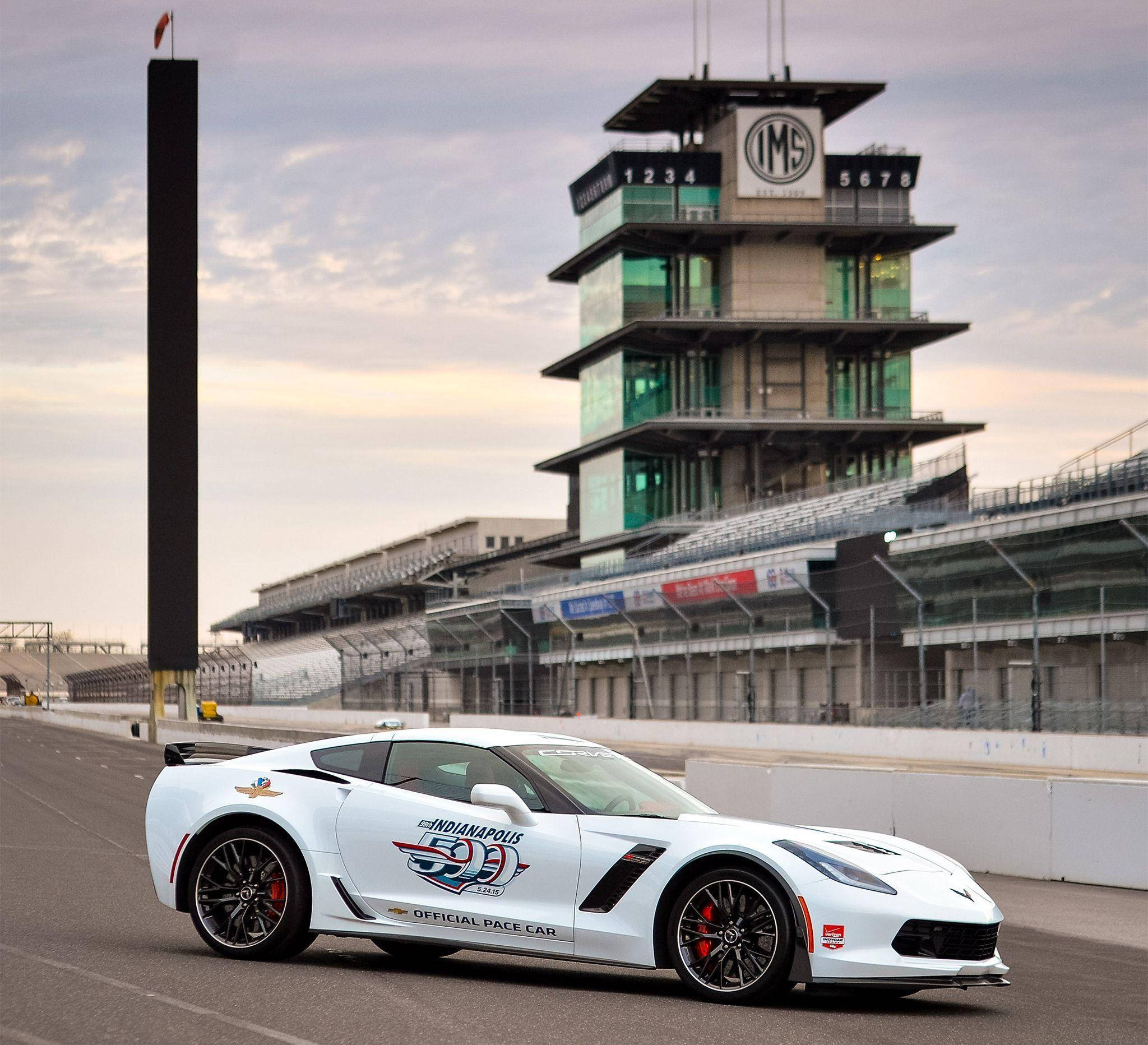 印第安納波利斯500賽道白色跑車 壁紙 — 一輛帶有Indy 500貼紙的白色Corvette Pace Car停在空蕩蕩的賽道上，背景是陰天和標誌性的IMS塔。