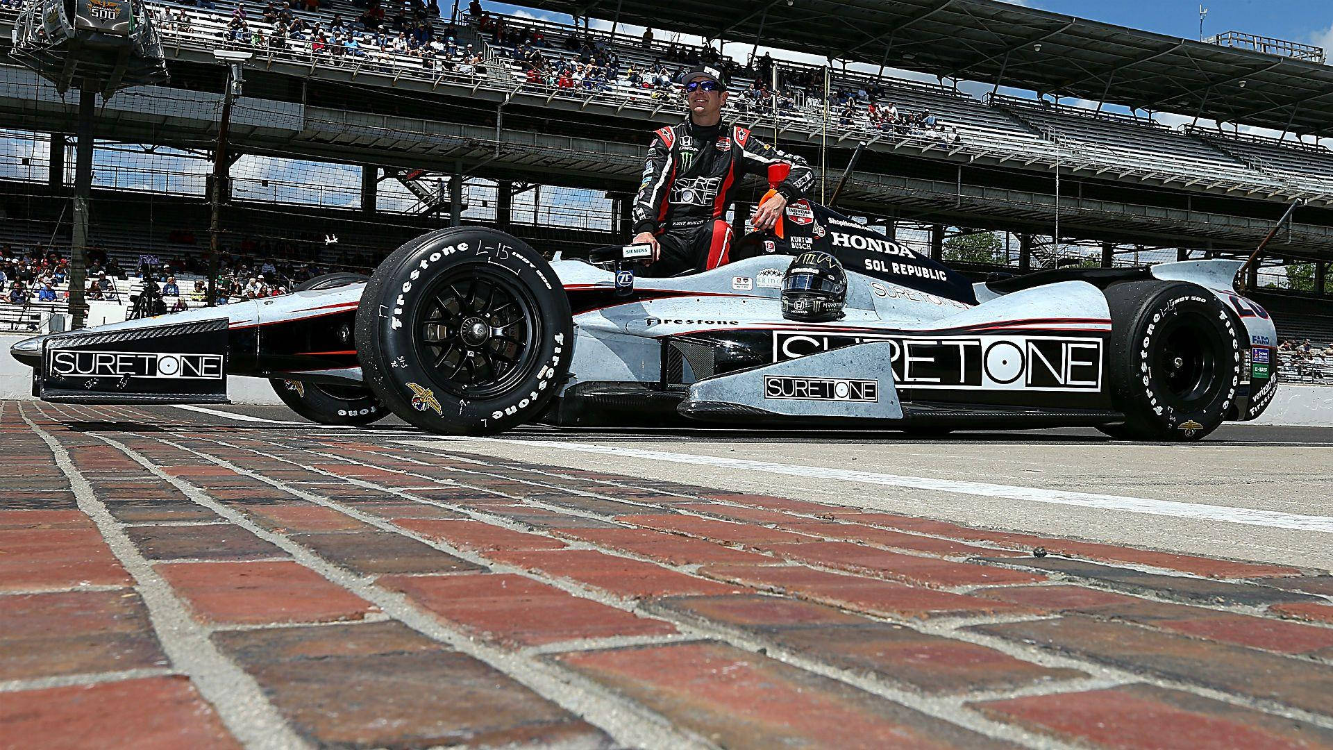 印第安納波利斯500本田F1賽車 壁紙 — 一輛帶有'SURETONE'品牌的時尚銀黑色開輪賽車停在紅磚維修區車道上，在明亮的日光下，附近有一位穿著裝備的車手。