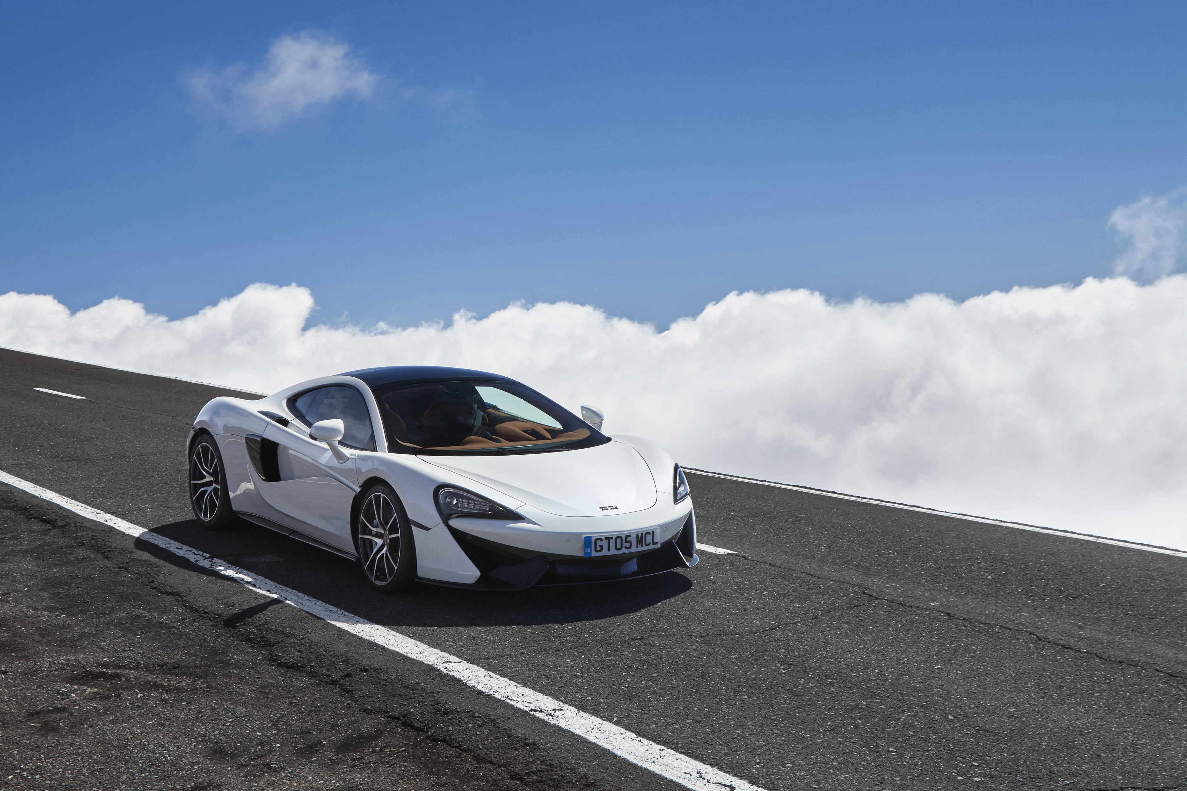 白色麥拉倫570 GT 壁紙 — 一架時尚的白色McLaren跑車在晴朗藍天下，行駛在雲海之上的高山公路上。