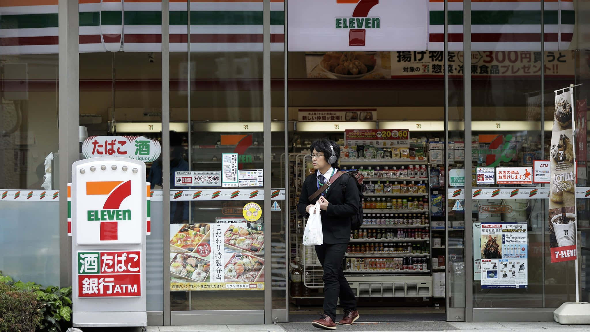 旅途中的7-Eleven，滿足你的零食與飲品需求 壁紙 — 一個人戴著耳機走過一間燈火通明的7-Eleven店舖，店舖有日文招牌和玻璃門反射街道。