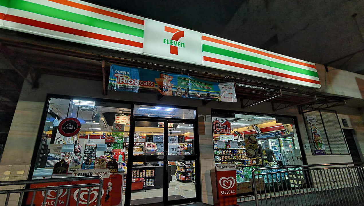 7-Eleven，享受冰涼美味 壁紙 — 夜晚燈火通明的7-Eleven店舖，色彩繽紛的招牌和玻璃門展示內部貨架上的商品。