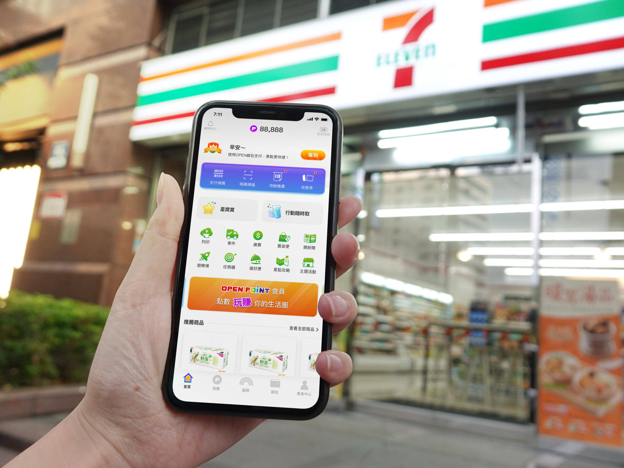 7-Eleven CLiQQ應用程式 壁紙 — 一隻手拿著智能手機，在夜晚燈光明亮的便利店前顯示7-Eleven應用程式介面。